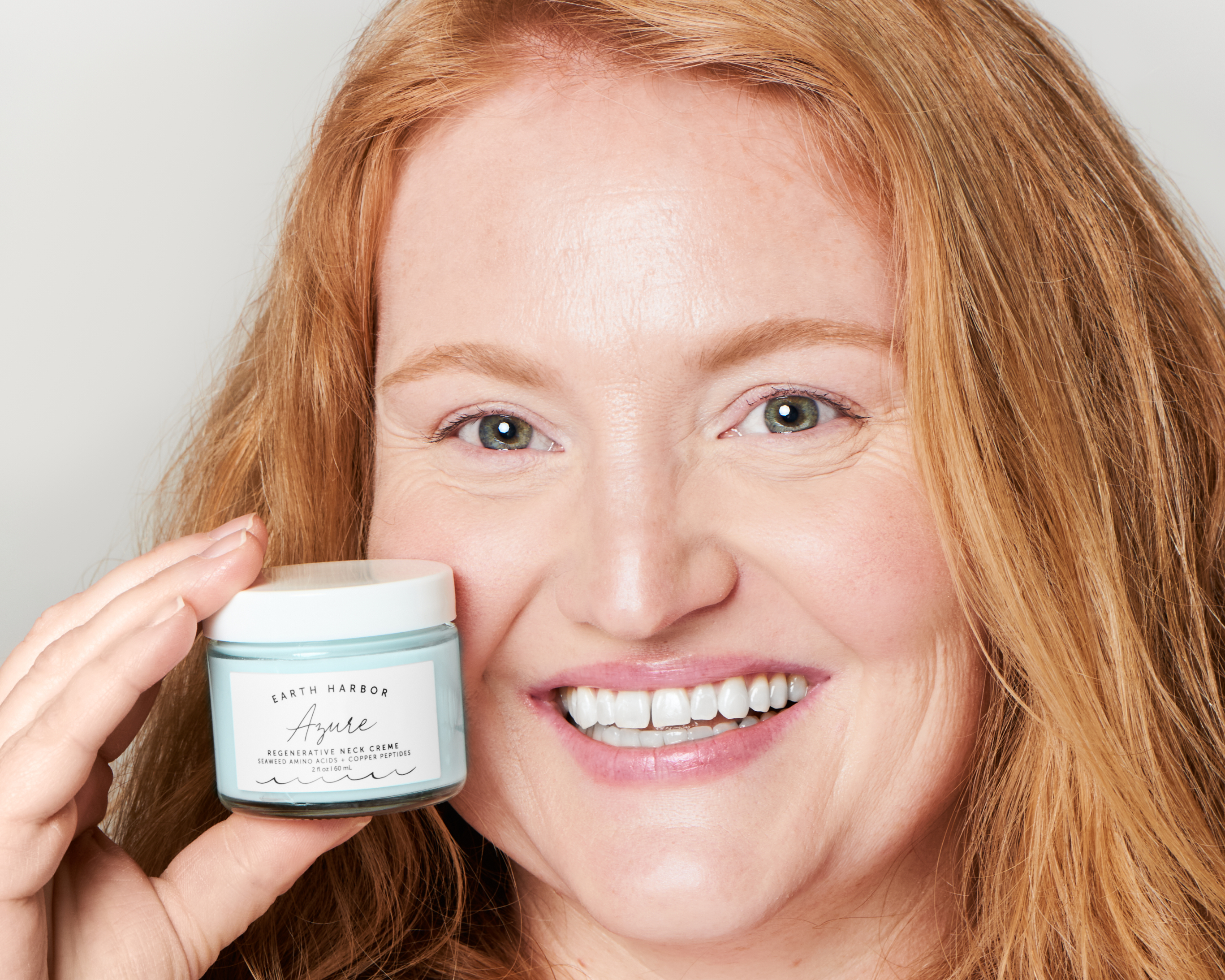 Smiling woman holding a small skincare
jar next to her face against a neutral background.​
