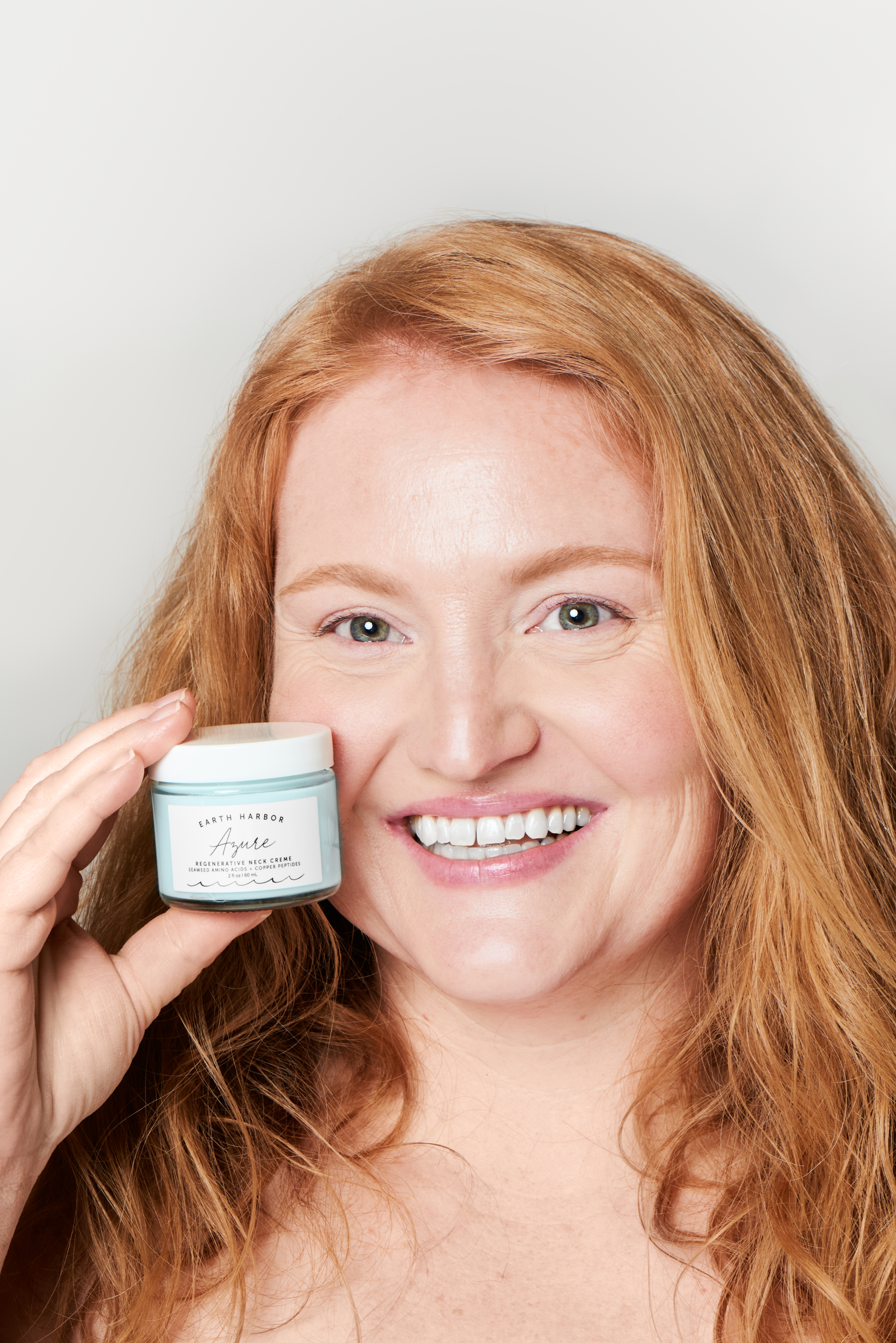 Smiling woman holding a small skincare
jar next to her face against a neutral background.​