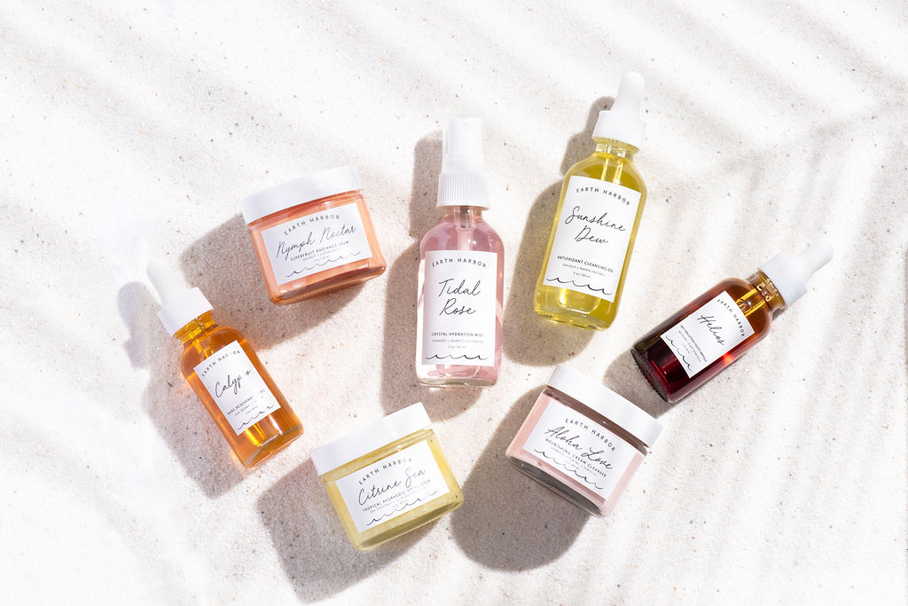 Assorted Earth Harbor skincare bottles and jars arranged flat on white sand under
soft shadows.​