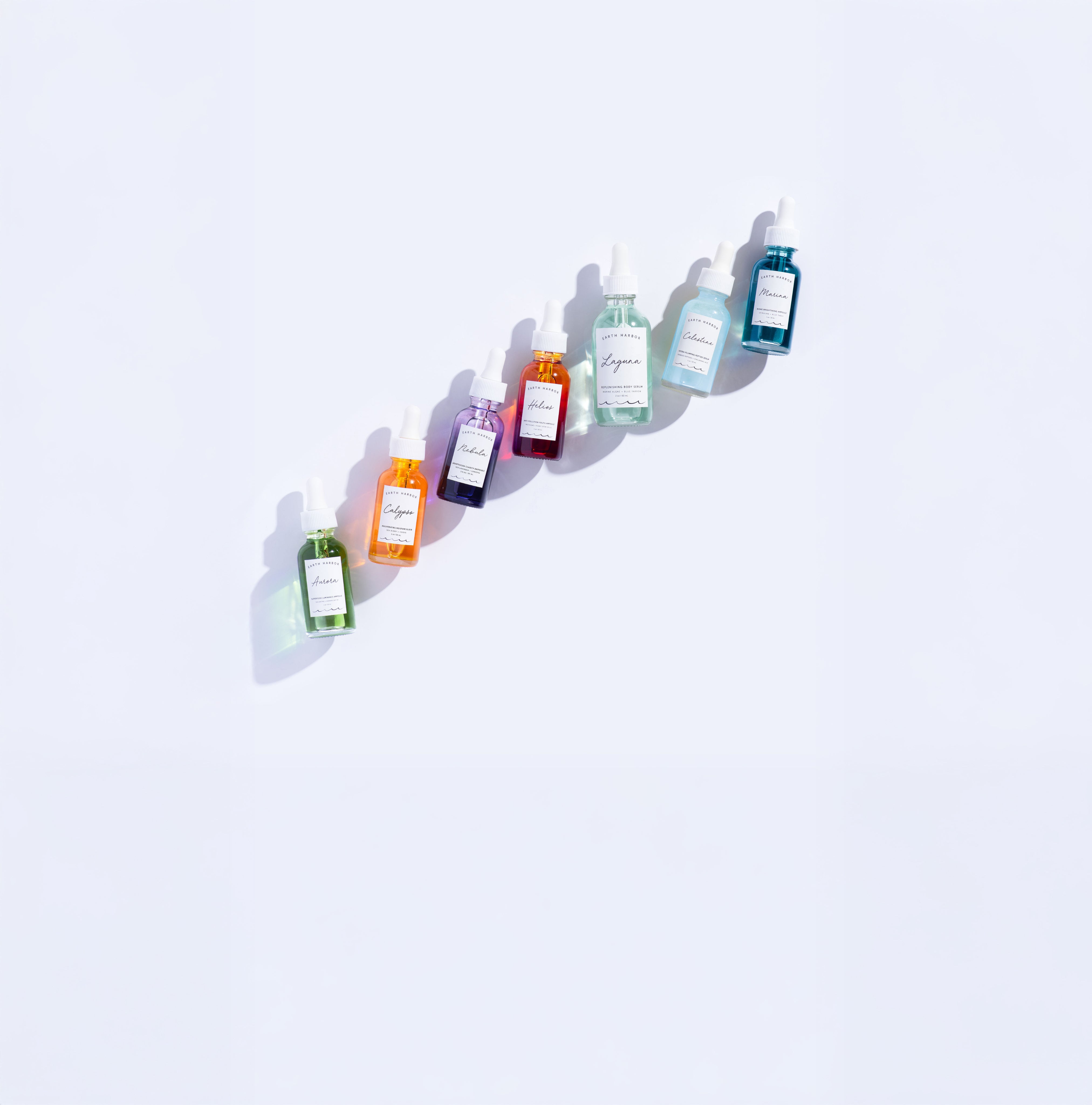 Colorful lineup of small Earth Harbor
serum bottles arranged diagonally on a light
background.​