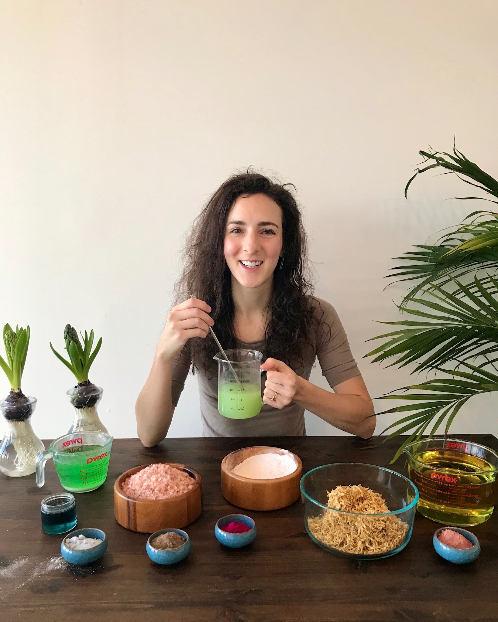 Woman mixing green skincare ingredients
at a table with bowls and plants.​