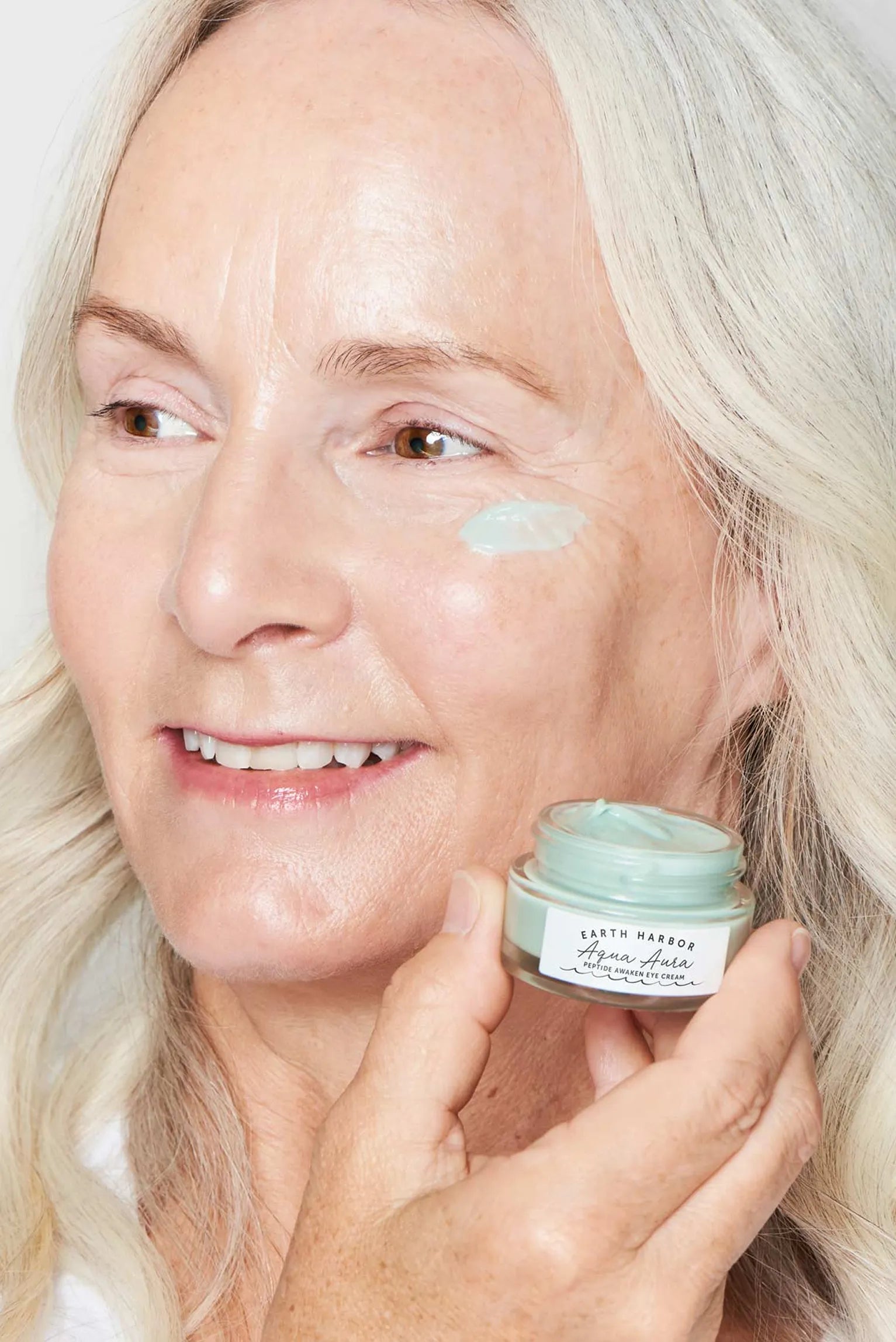 Smiling woman with grey hair
holding an Earth Harbor eye
cream jar with product
dabbed under her eye.​