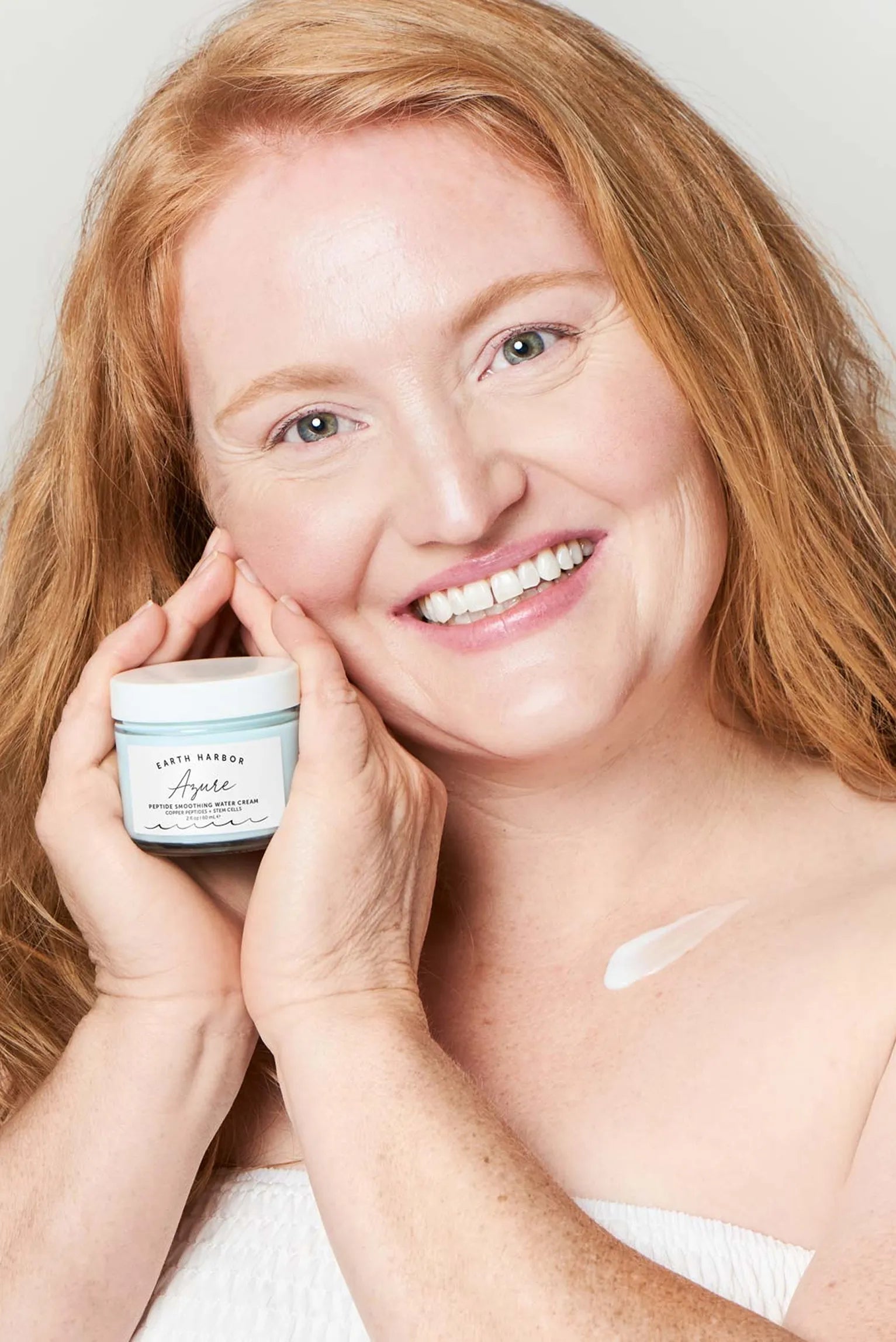 Smiling red-haired woman holding a jar of
Earth Harbor Azure cream with a dab of
lotion on her collarbone.​