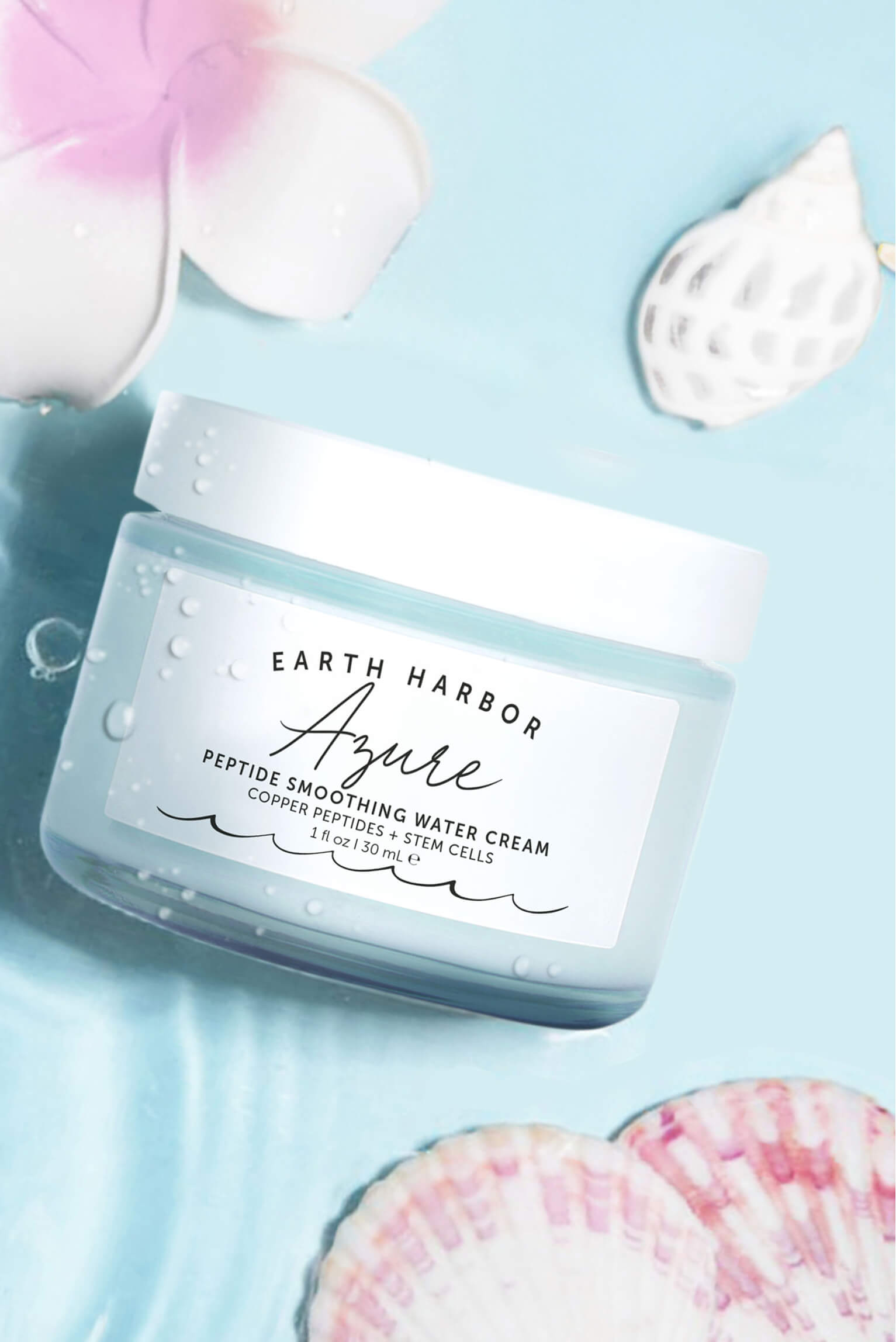 Earth Harbor Azure cream jar resting in
clear water next to seashells and a pink
tropical flower.​
