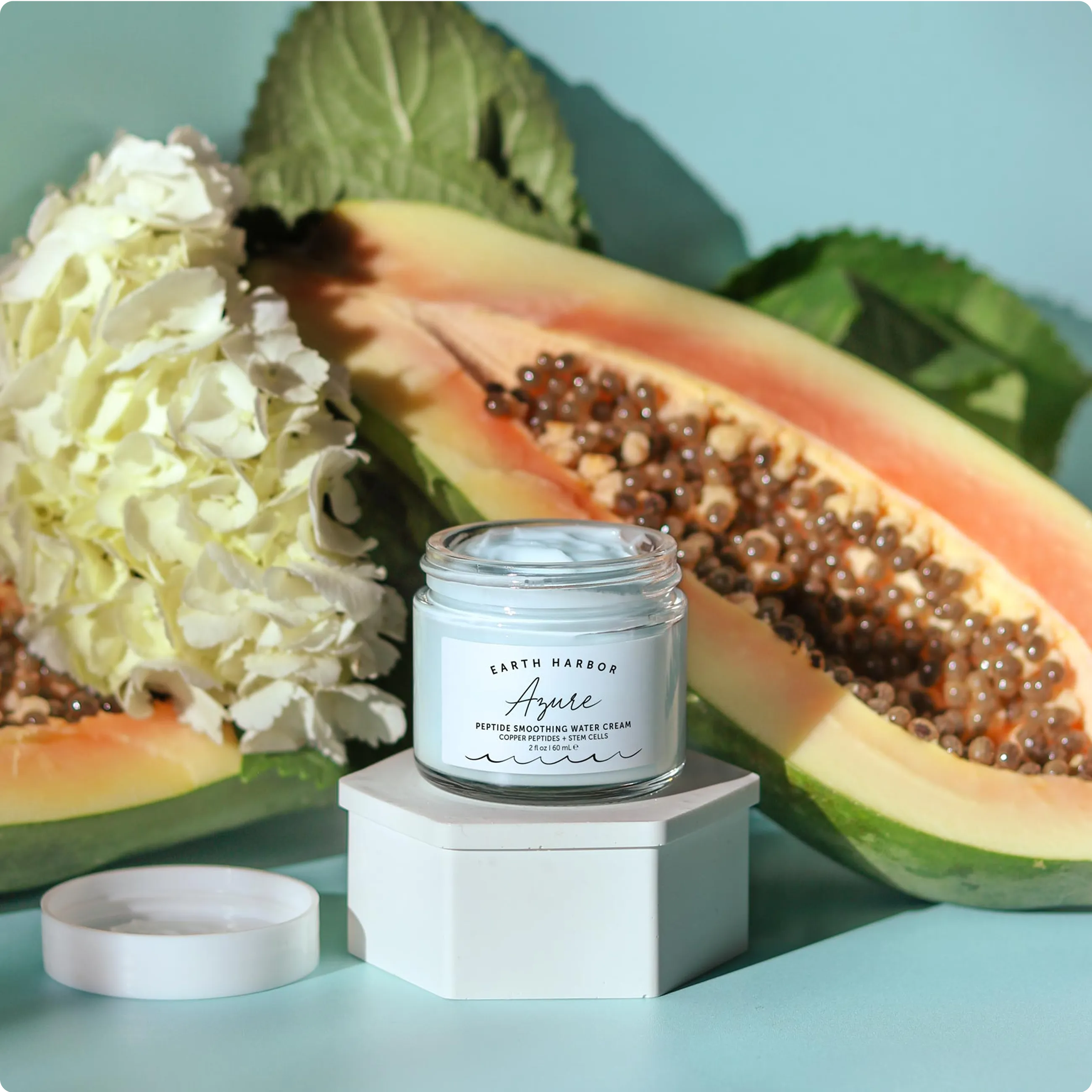 Open jar of Earth Harbor Azure cream on a
white block with a sliced papaya and white
flowers behind it.​