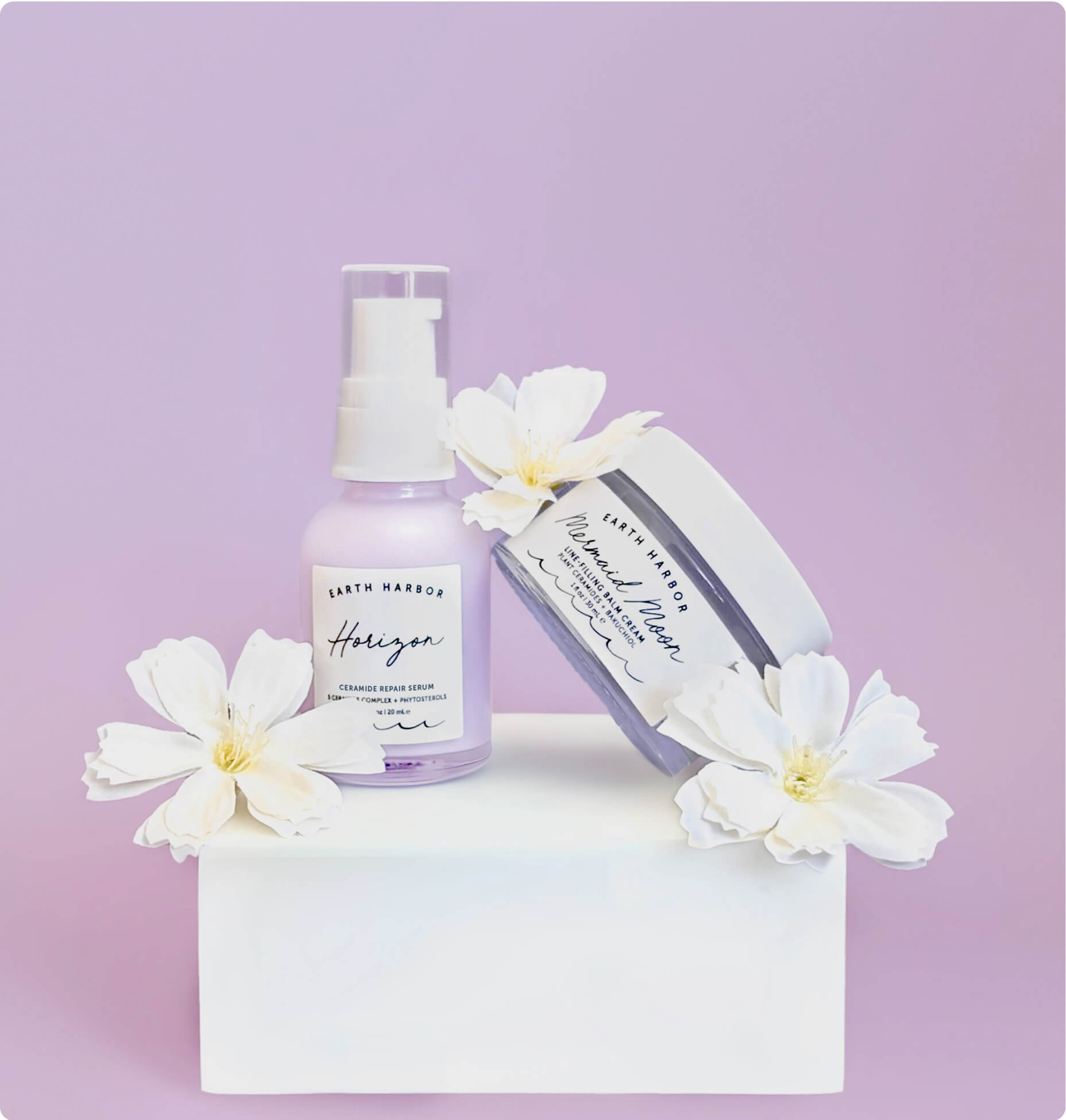Earth Harbor Horizon pump bottle and Mermaid Moon jar on a white stand with white flowers
against a lilac background.​