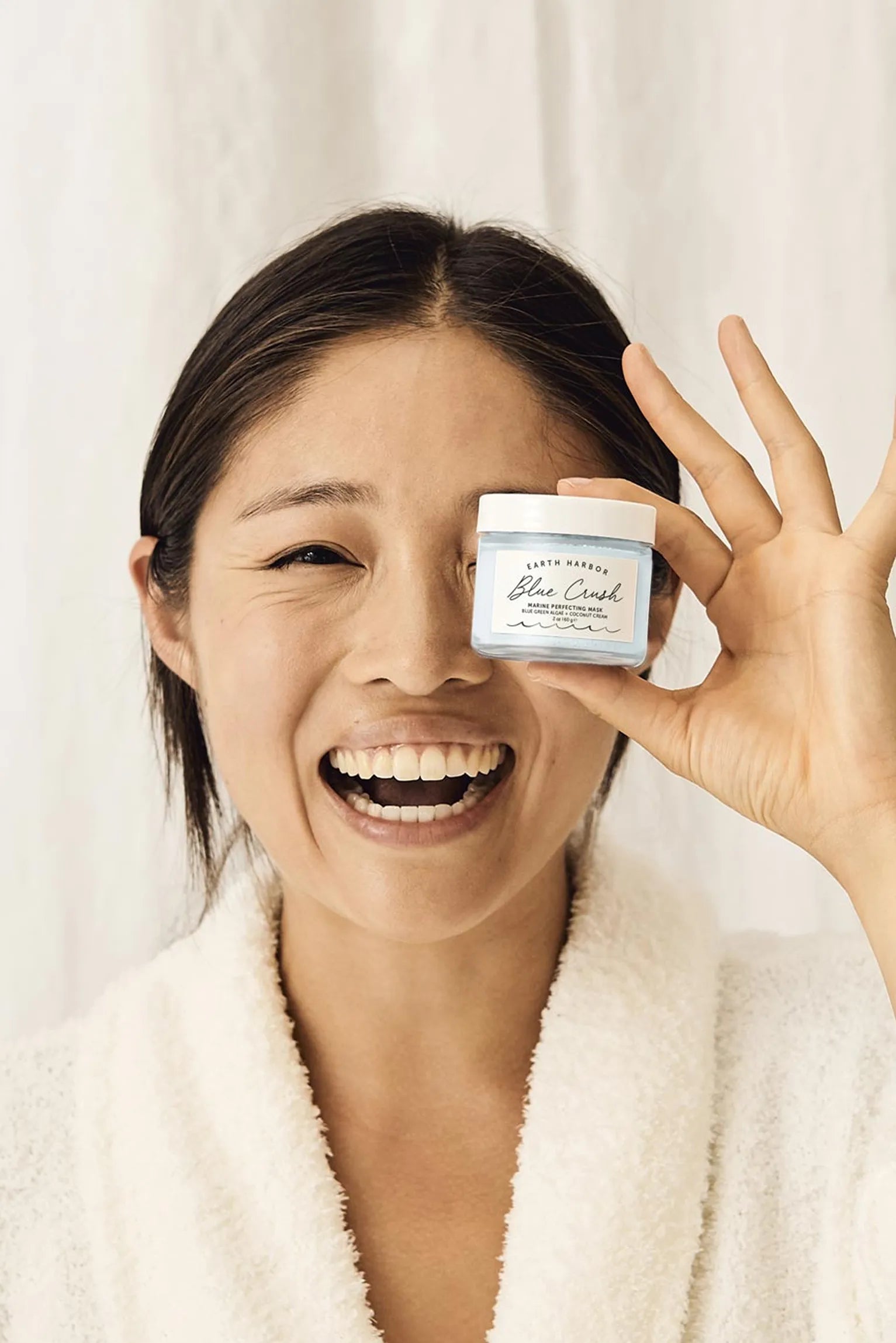 Laughing woman in a white robe holding a
jar of Earth Harbor Blue Crush mask over
her eye.​