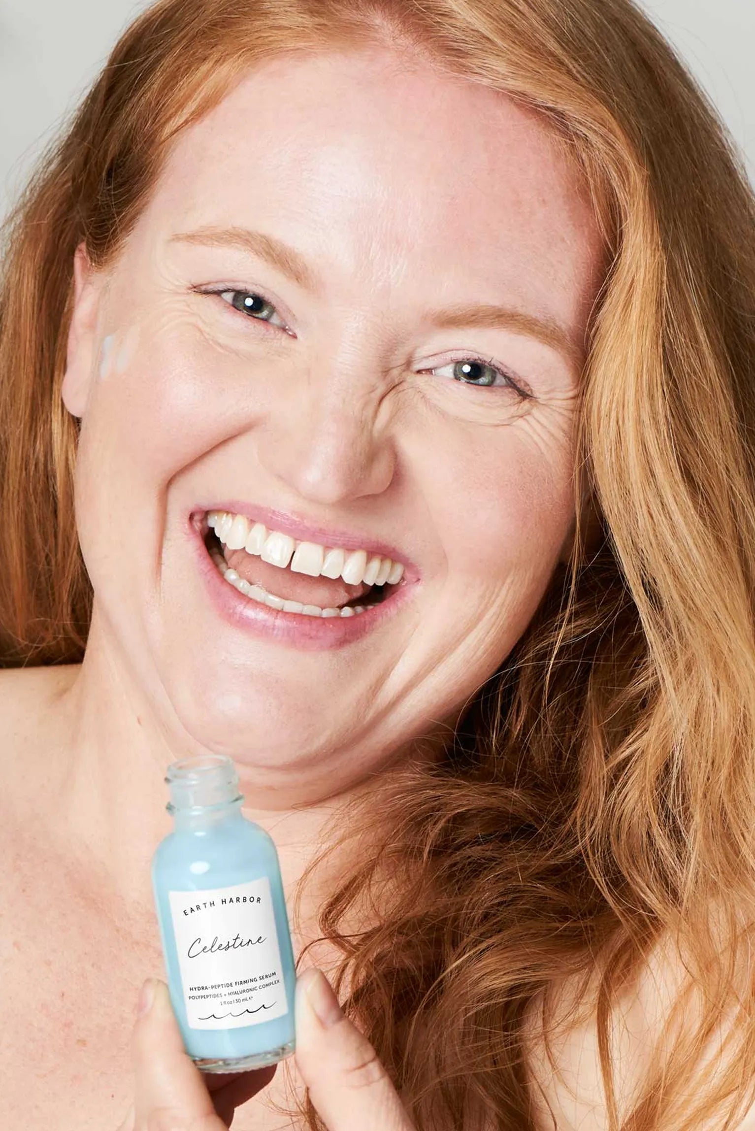 Laughing redhead woman holding a blue
bottle of Earth Harbor Celestine serum.​
