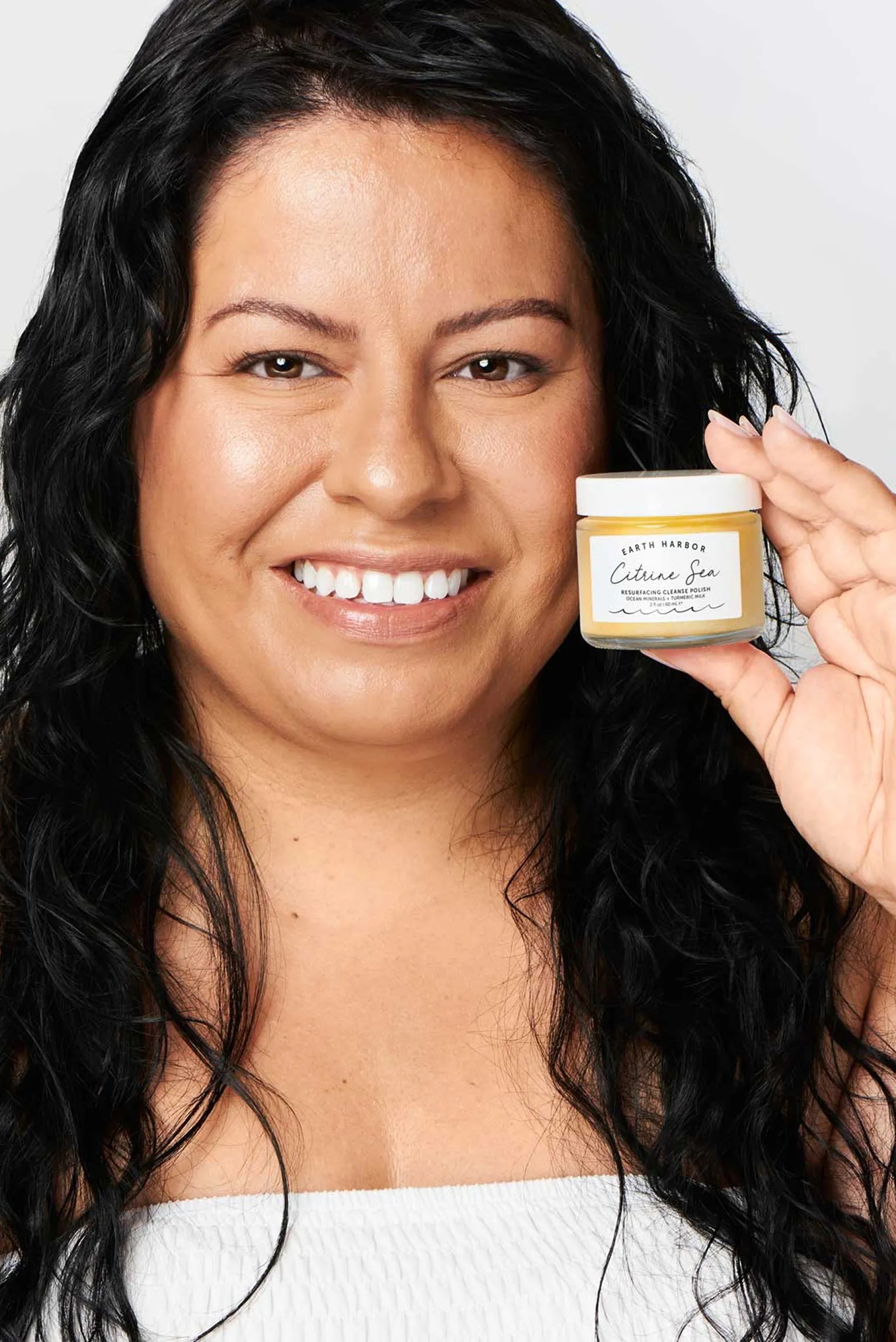 Smiling woman with wavy brown hair
holding a coral jar of Earth Harbor Nymph
Nectar balm next to her face.​