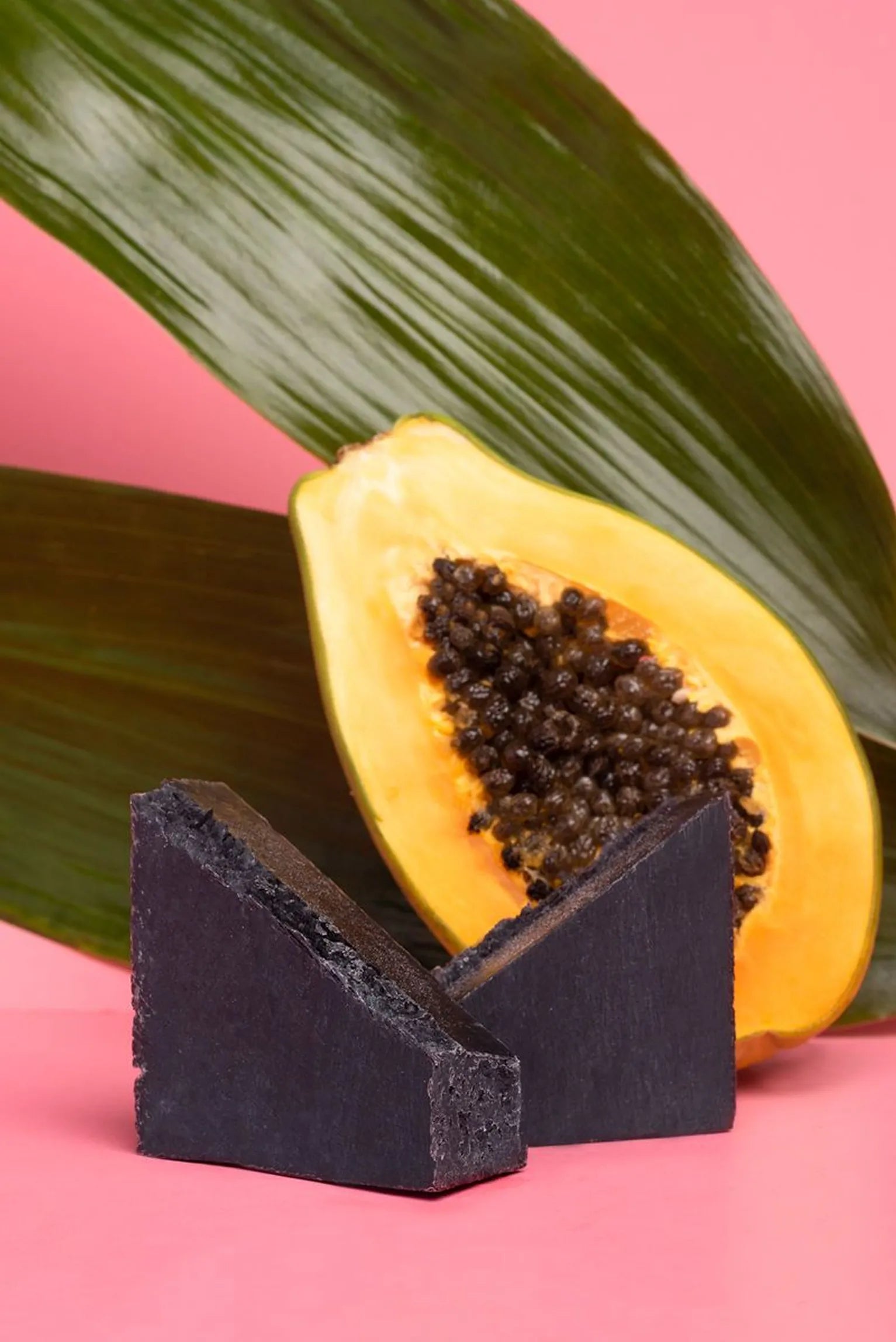 Two triangular wedges of black charcoal
soap in front of a sliced papaya fruit on a
pink backdrop.​