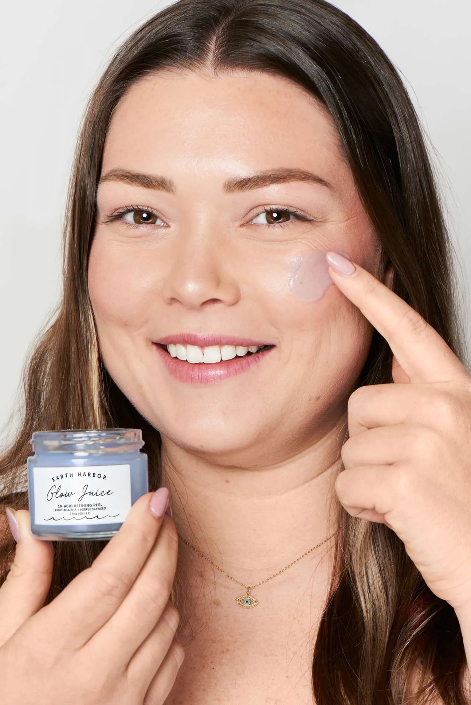 Smiling woman applying a purple gel mask
to her cheek while holding a jar of Earth
Harbor Glow Juice.​
