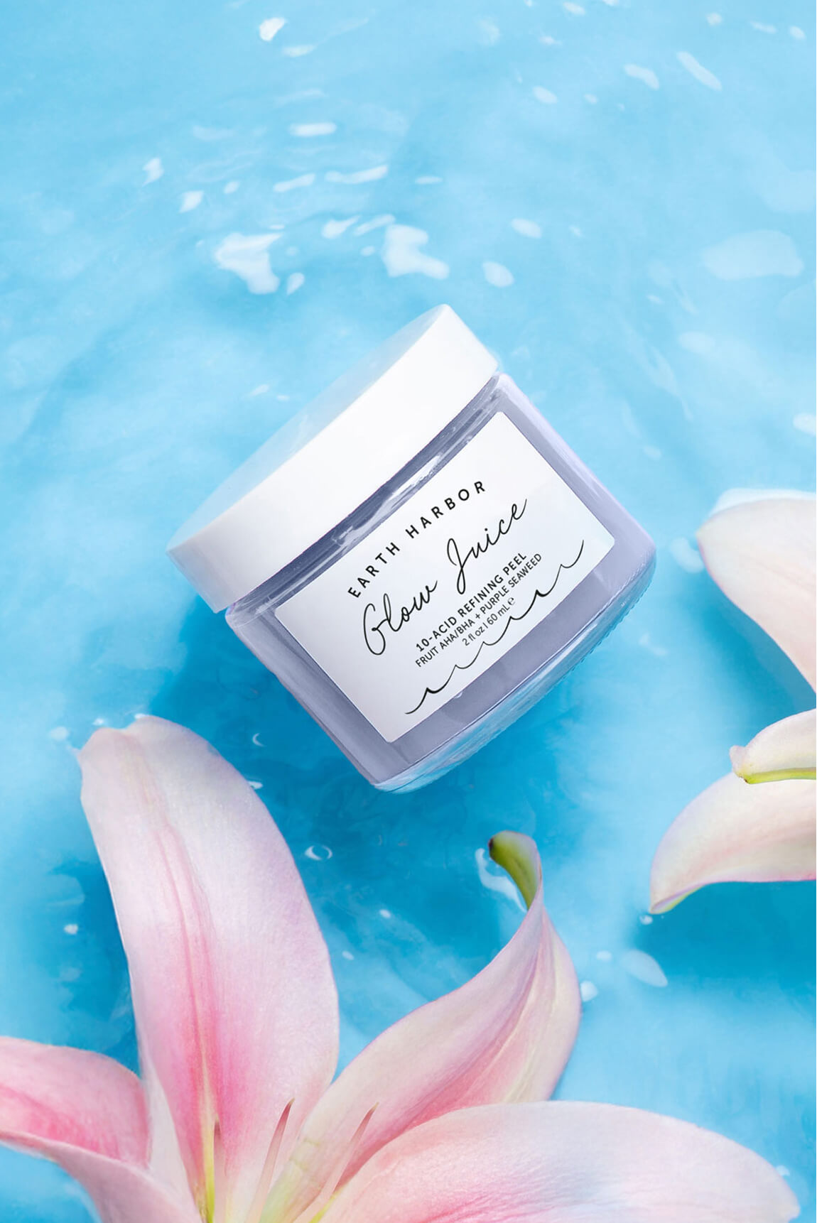 Jar of Earth Harbor Glow Juice mask in
clear, rippling blue water surrounded by
pink lily petals.​