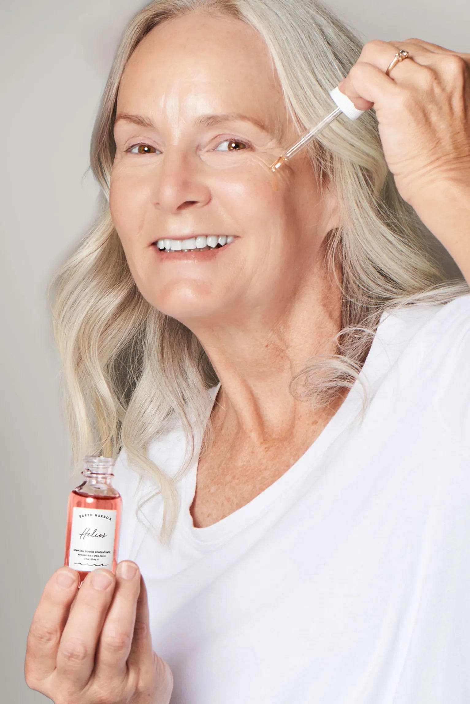 Smiling older woman with long silver hair
applying a drop of pink Earth Harbor Helios
concentrate to her face.​