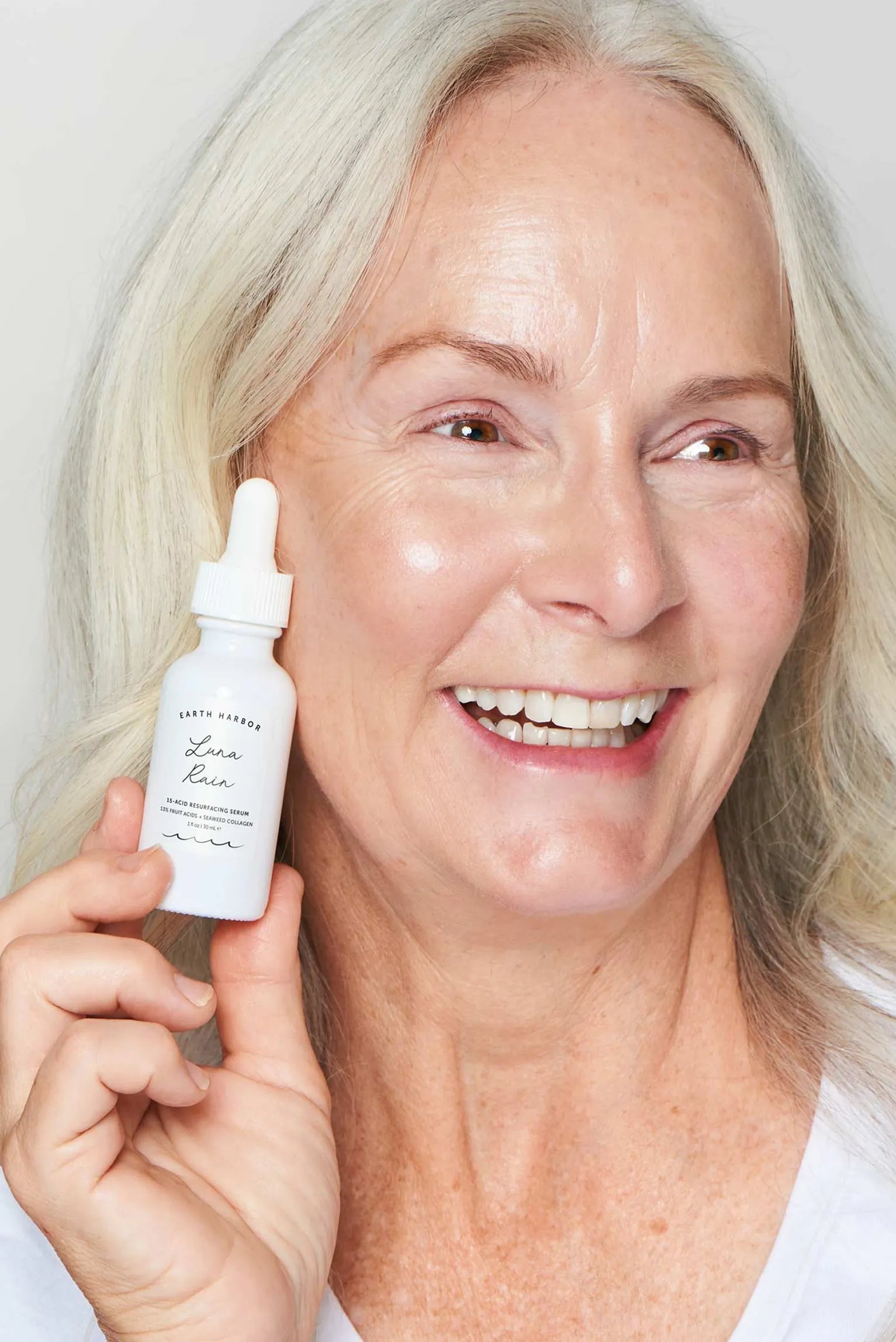 Smiling mature woman with white hair
holding a white Earth Harbor Luna Rain
dropper bottle near her cheek.​