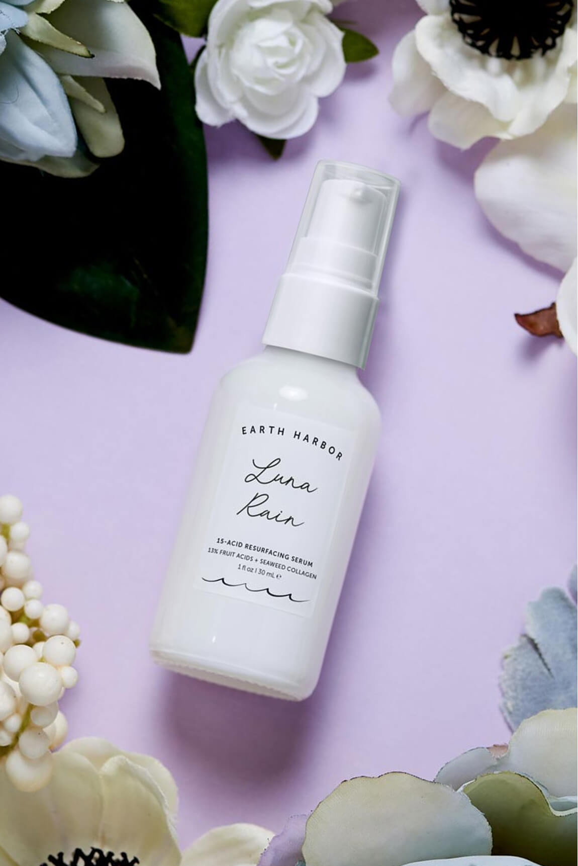 White bottle of Earth Harbor Luna Rain
serum lying flat on a purple background
surrounded by white flowers.​