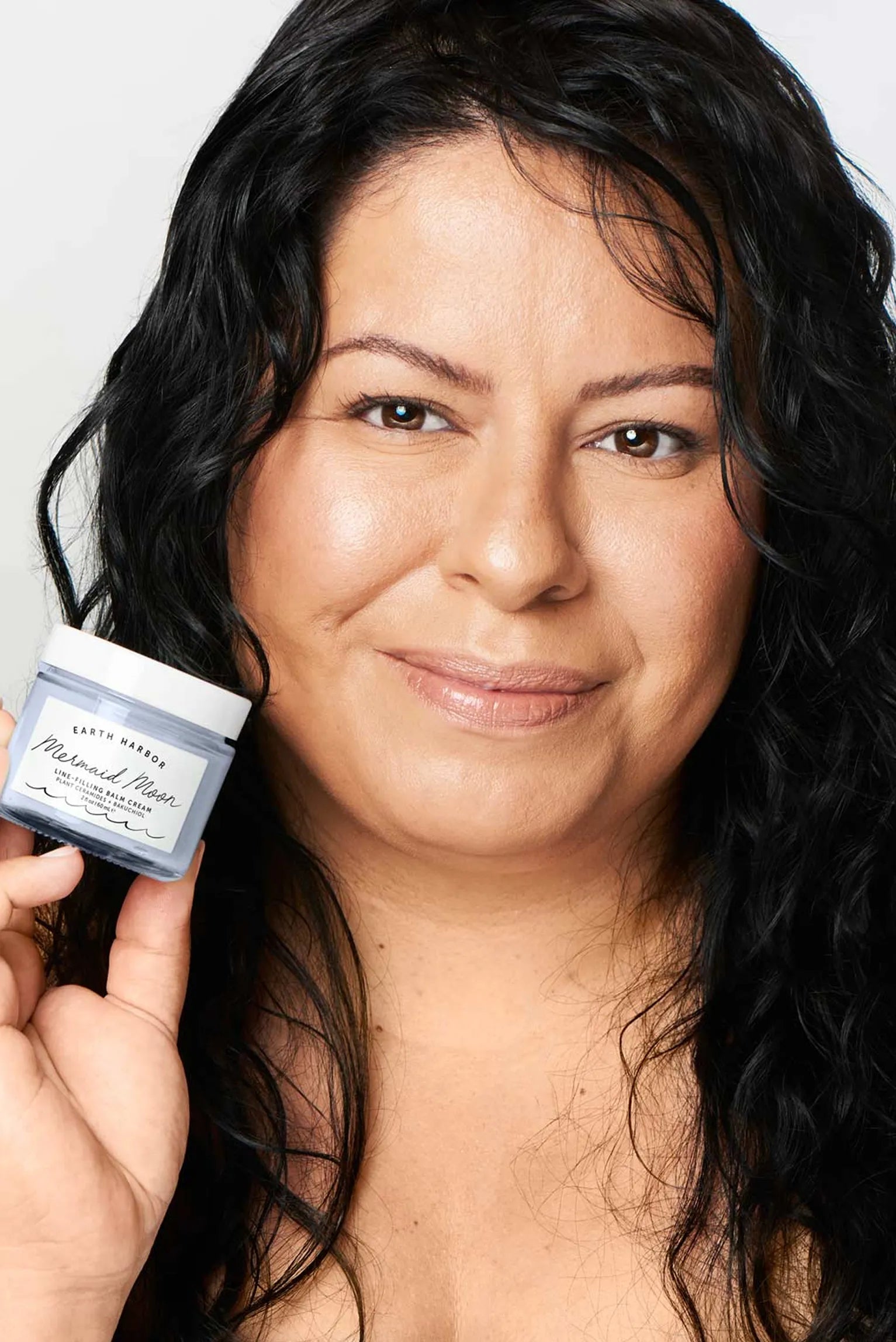 Smiling woman with long, dark wavy hair
holding a small purple jar of Earth Harbor
Mermaid Moon cream.​