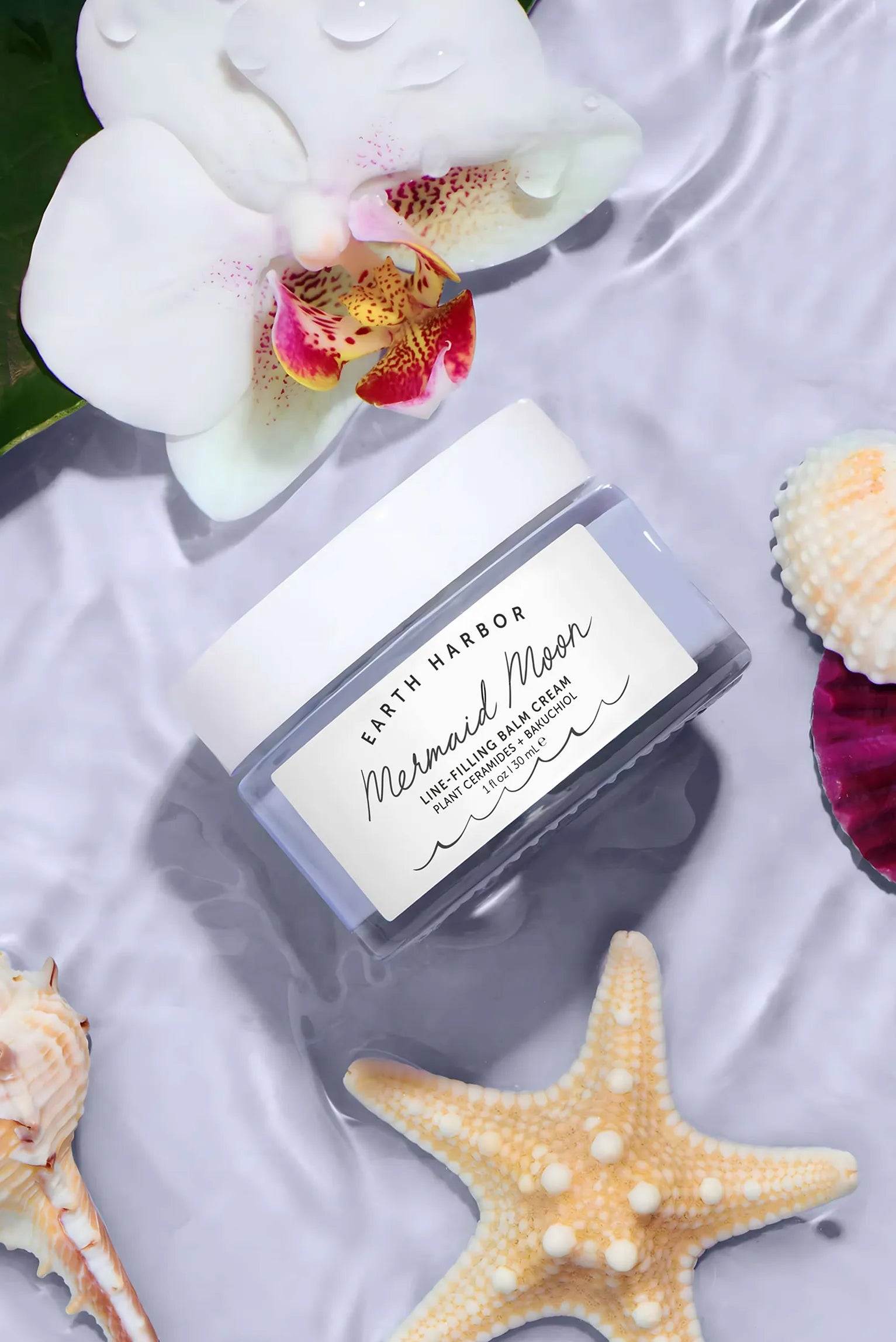 Earth Harbor Mermaid Moon jar floating in
rippled water next to a white orchid, a
starfish, and seashells.​