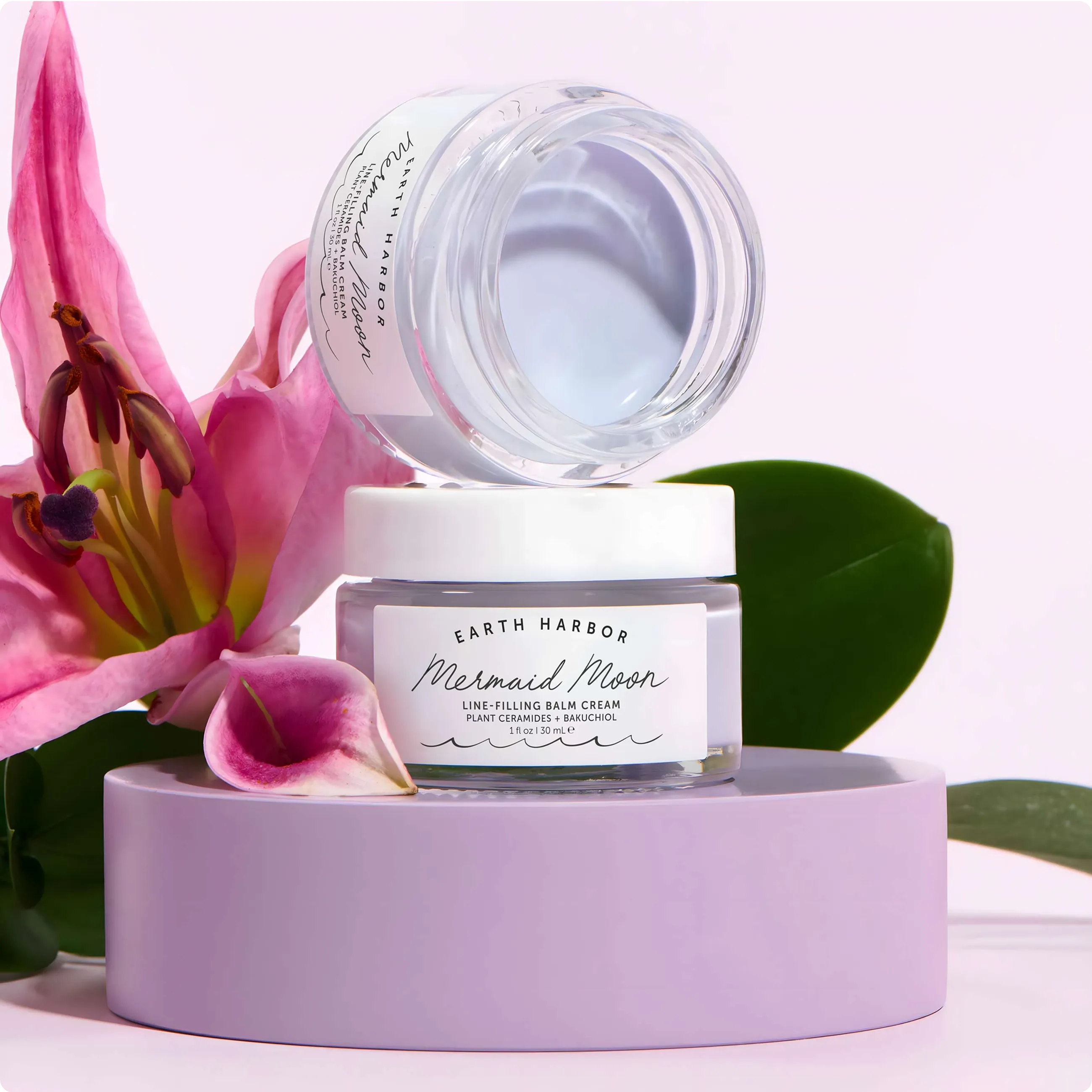 Earth Harbor Mermaid Moon cream jar
stacked on a purple platform next to a
vibrant pink lily flower.​