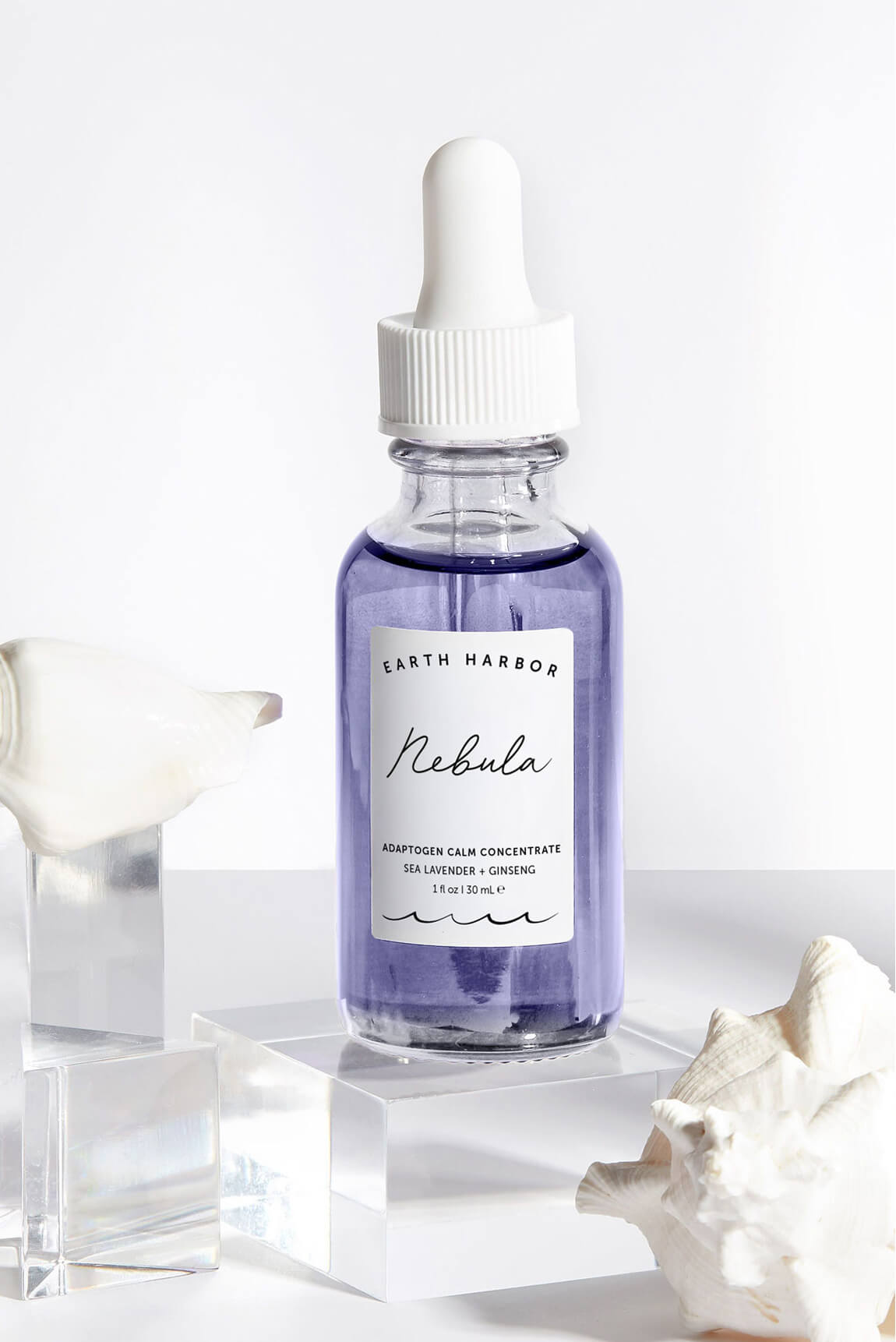 Earth Harbor Nebula ampoule bottle
resting on a clear acrylic block next to a
white seashell.​