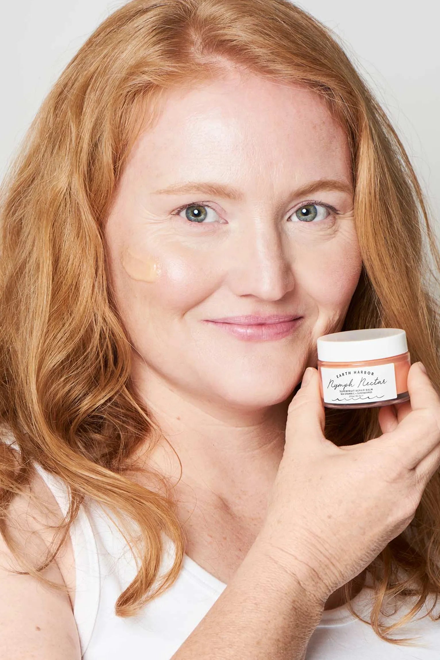 Smiling woman with strawberry blonde hair
holding a small Earth Harbor skincare jar by her
face.​