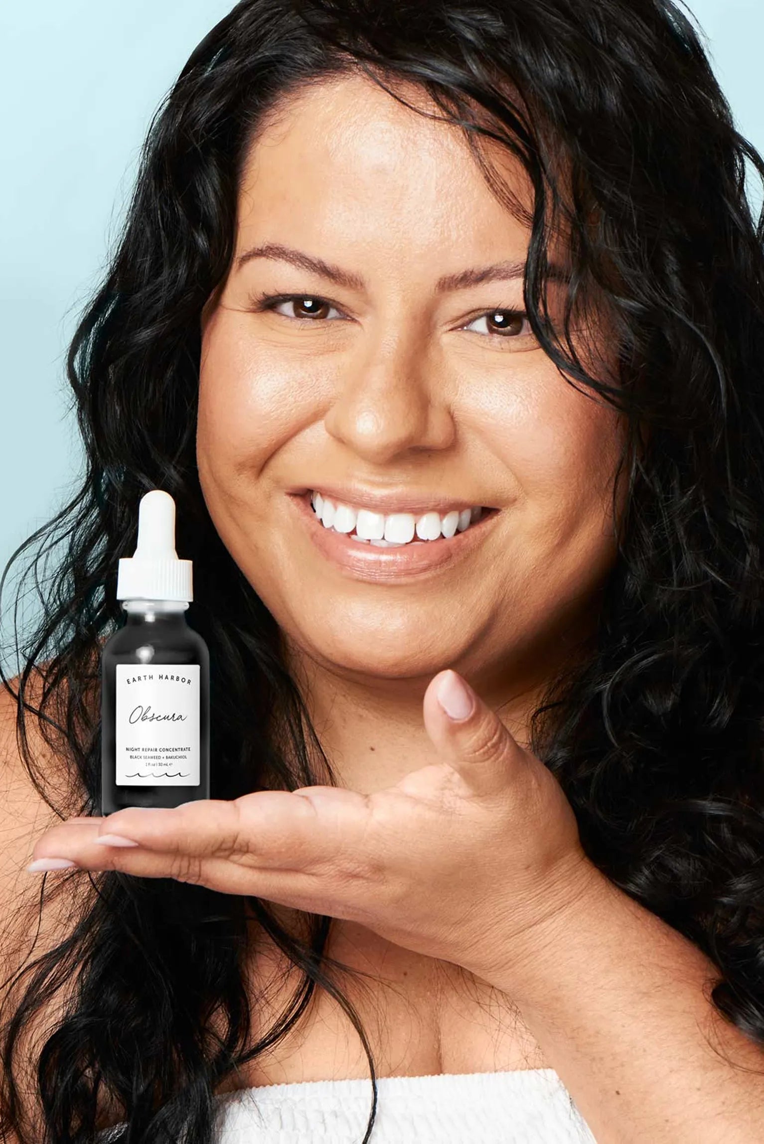 Smiling woman with long, dark wavy hair
holding a bottle of Earth Harbor Obscura
serum in her open palm.​
