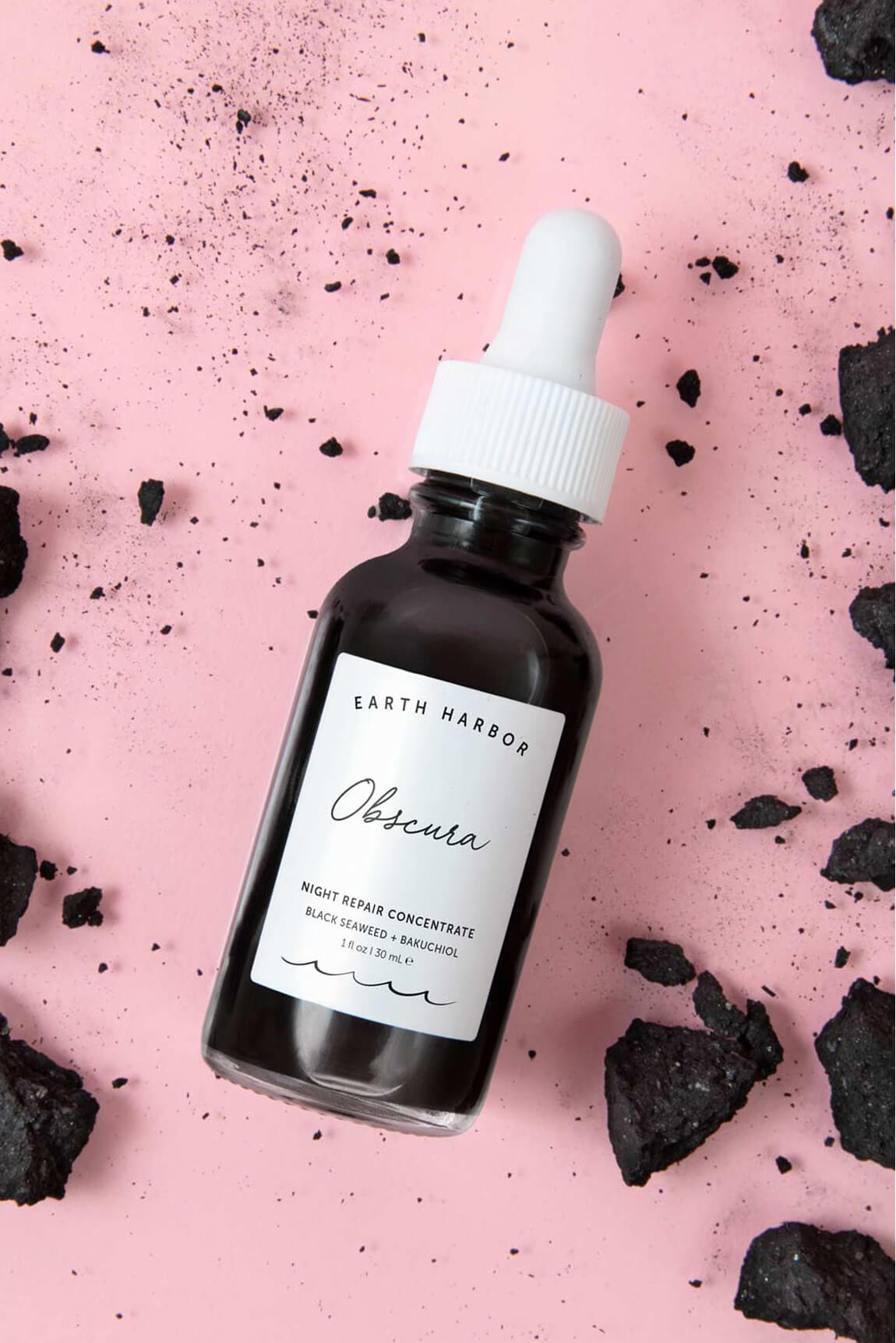 Earth Harbor Obscura dropper bottle lying
on a textured pink surface scattered with
chunks of black charcoal.​