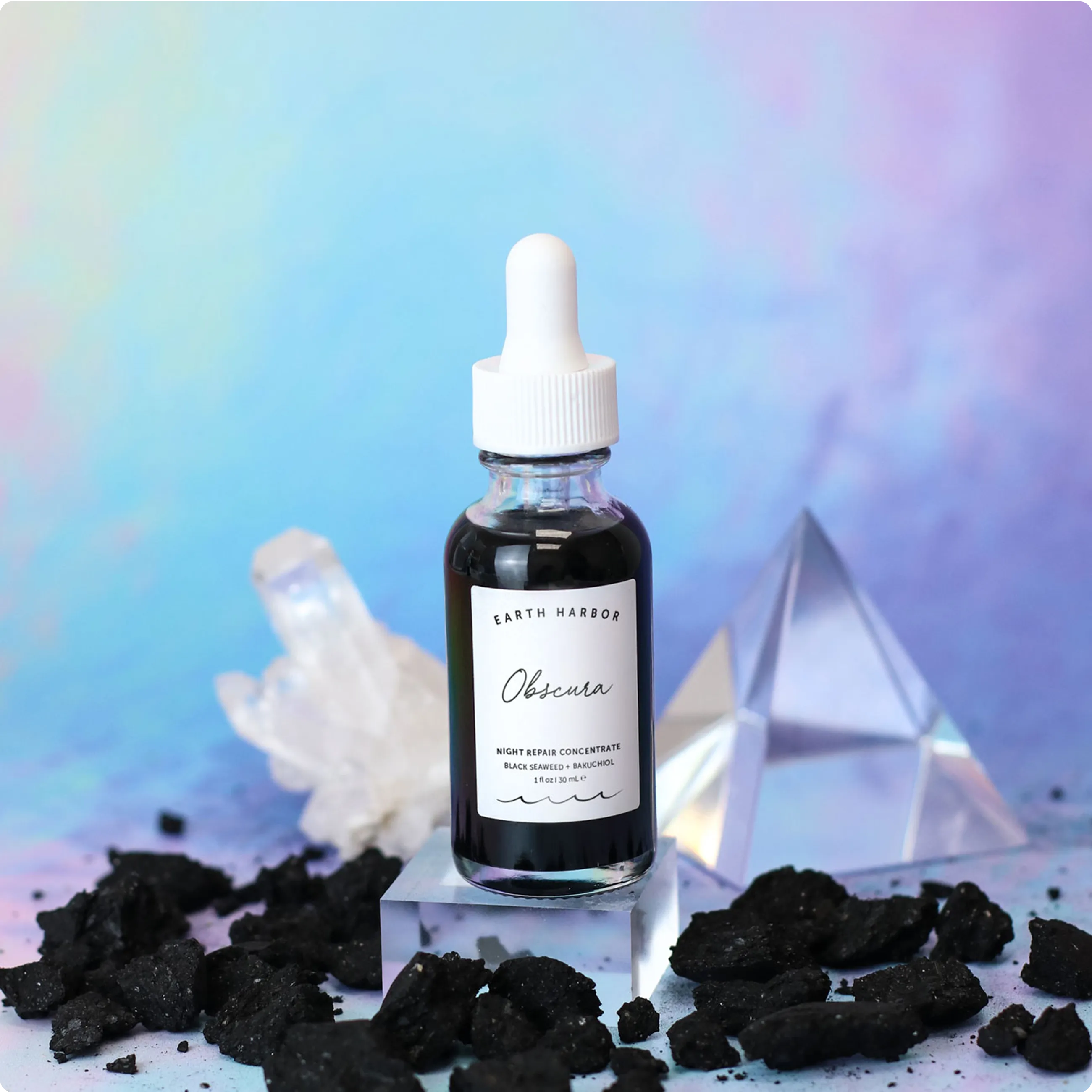 Earth Harbor Obscura dropper bottle on a
glass cube next to clear quartz crystals and
black charcoal pieces.​