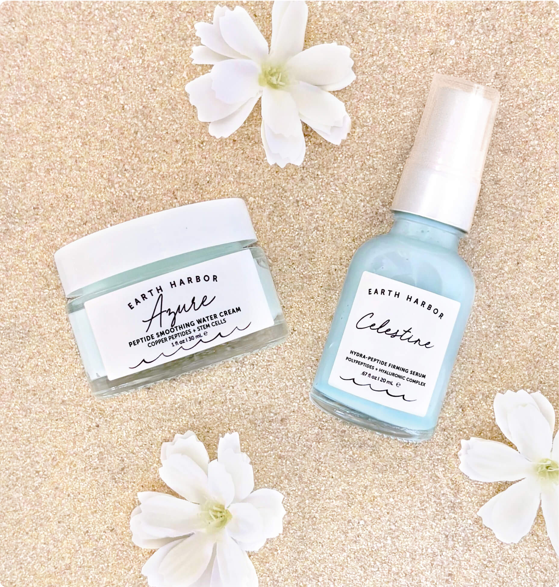 Earth Harbor Azure jar and Celestine pump bottle resting on sand next to small white flowers.​