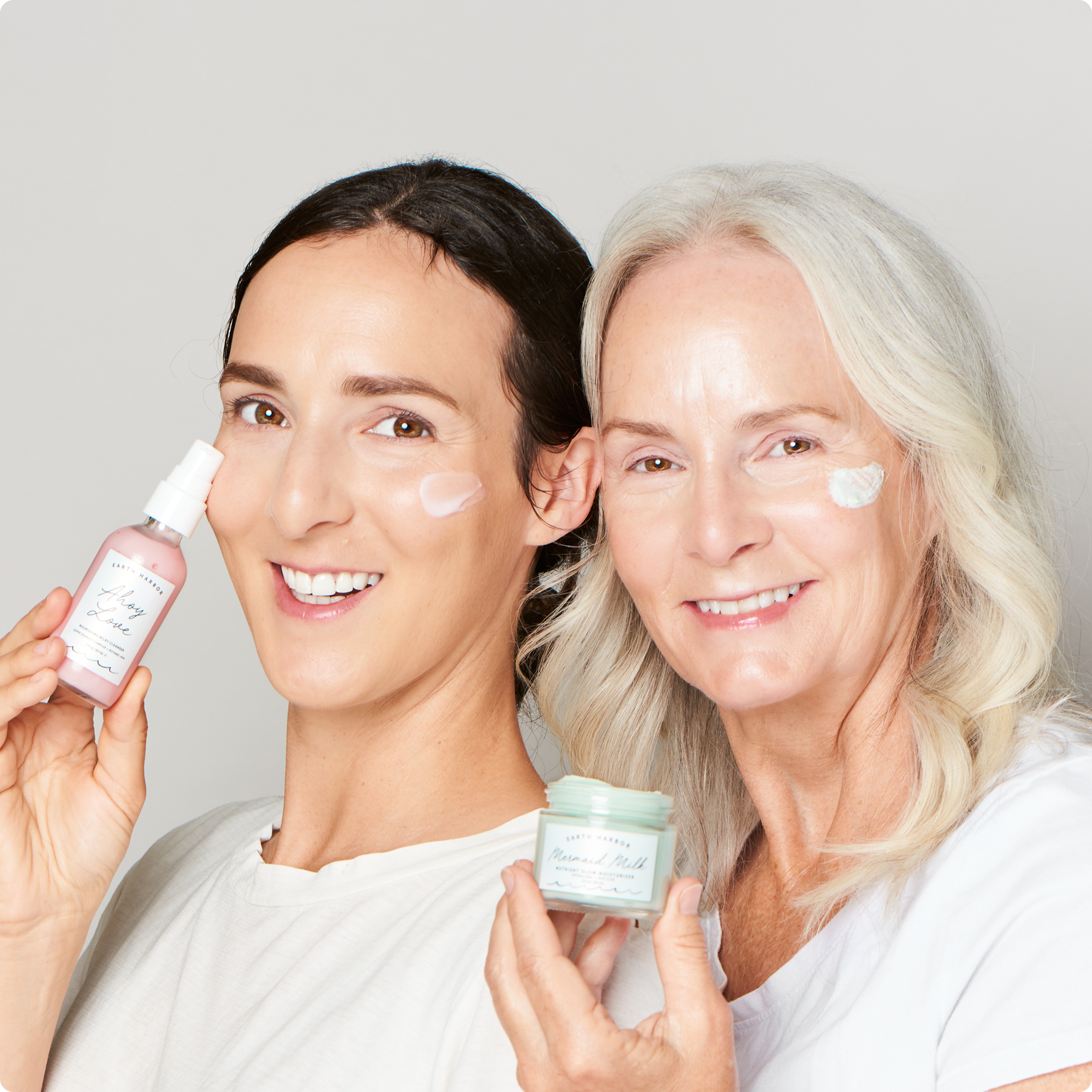 Two women smiling while holding
skincare products with cream applied to their cheeks.