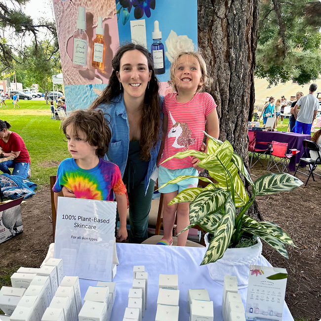 Woman and two children smiling at an
outdoor market booth with skincare products​