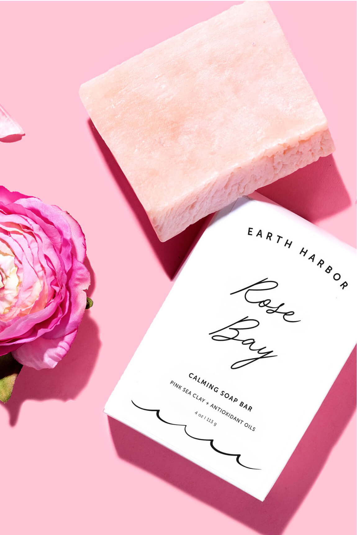 Earth Harbor Rose Bay soap bar and white
box packaging flat lay on a bright pink
surface with a pink flower.​