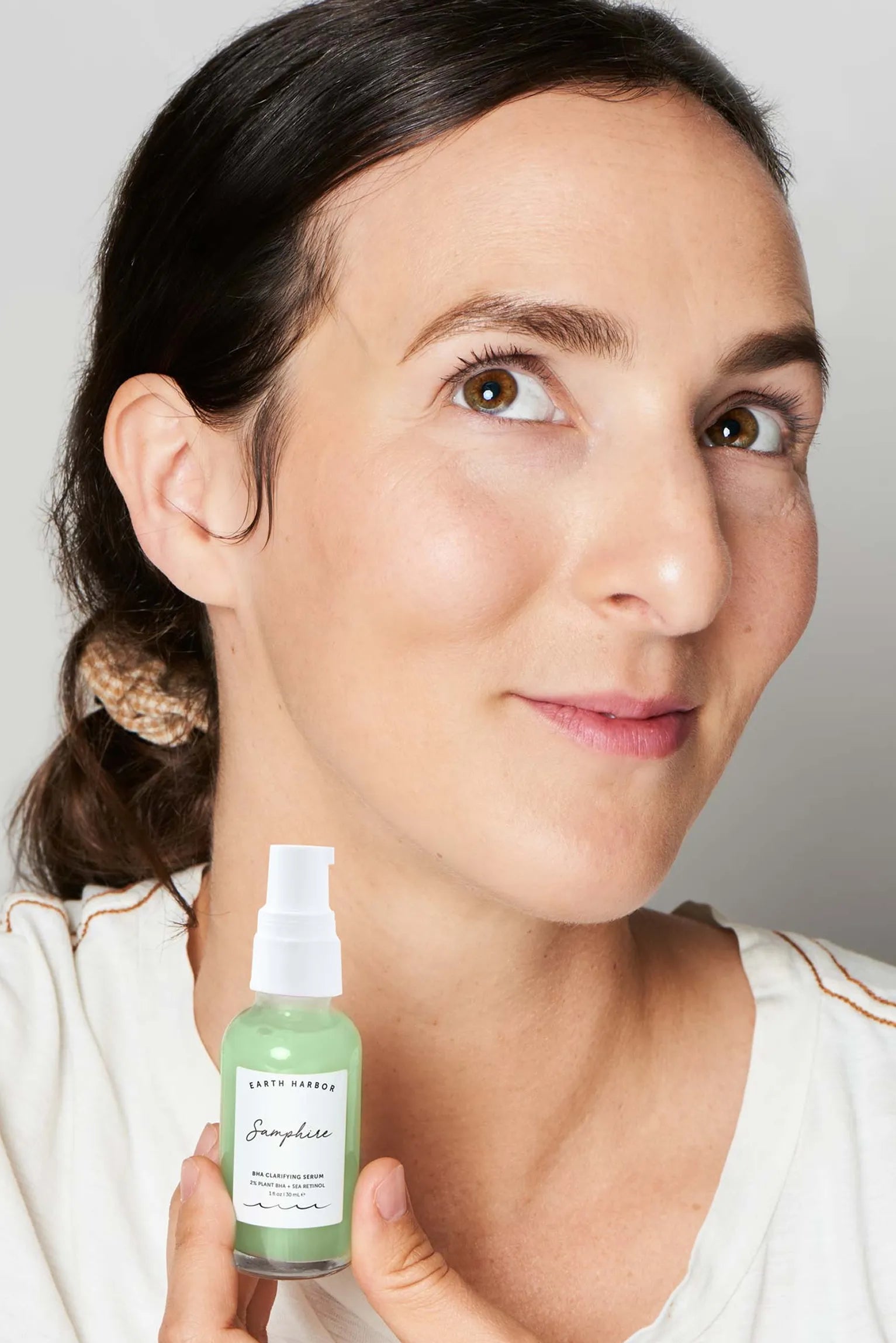 Woman holding a small green bottle of
Earth Harbor Samphire serum near her
chin.​