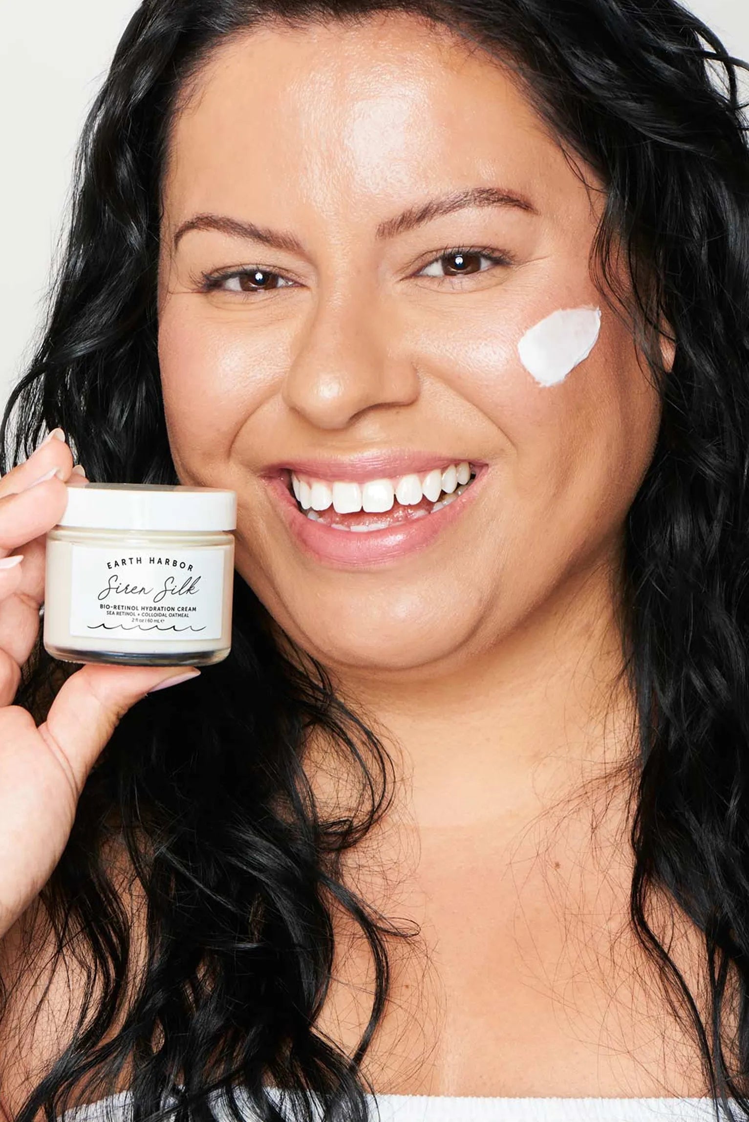 Smiling woman holding an Earth Harbor
Siren Silk jar with a dab of white face
cream on her cheek.​
