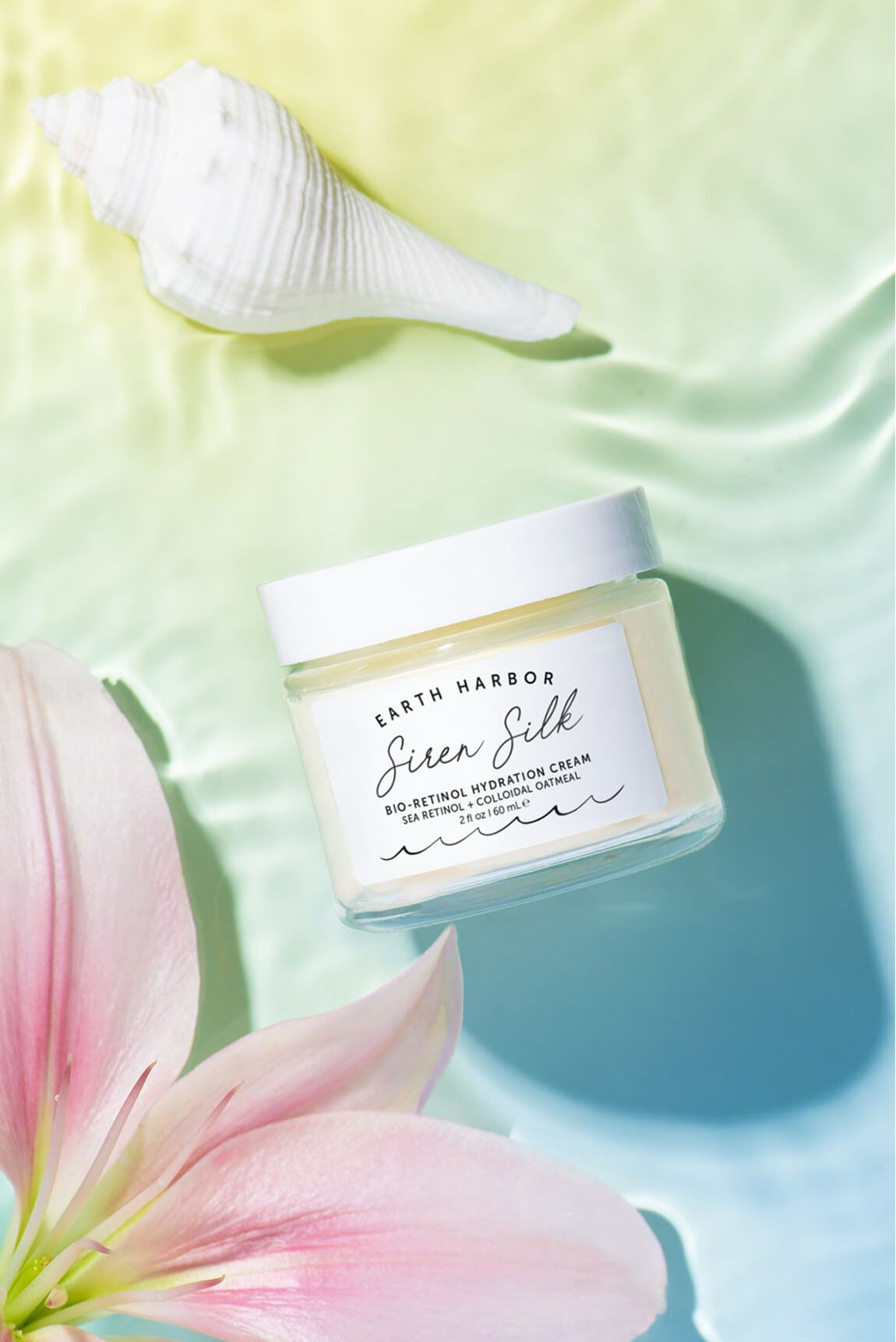 Earth Harbor Siren Silk jar on rippling
green water next to a pink lily flower and
white seashell.​