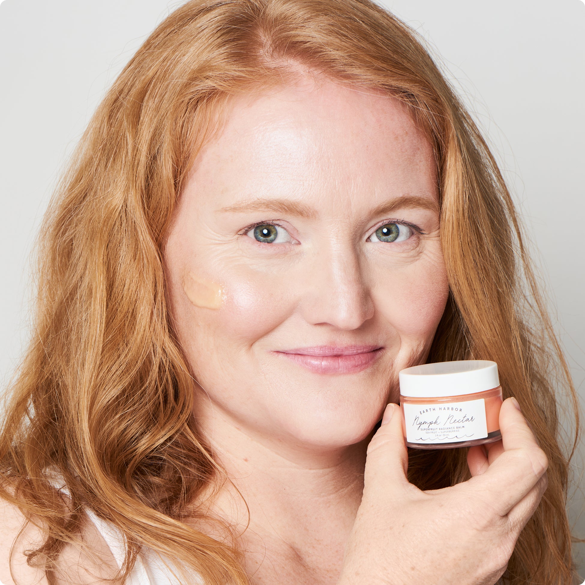 Woman with light skin smiling with a small
amount of cream applied to her cheek holding Nymph
Nectar​