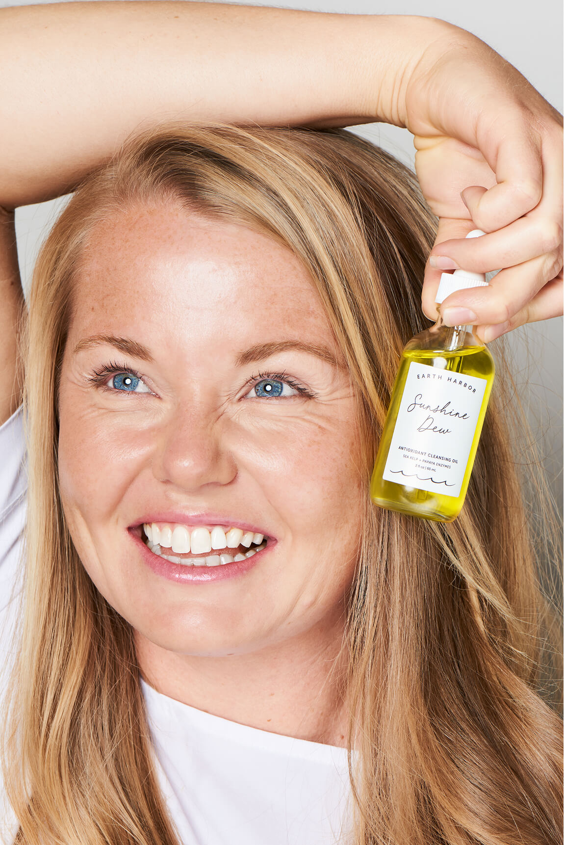 Smiling blonde woman holding an Earth
Harbor Sunshine Dew facial oil bottle next
to her face.​