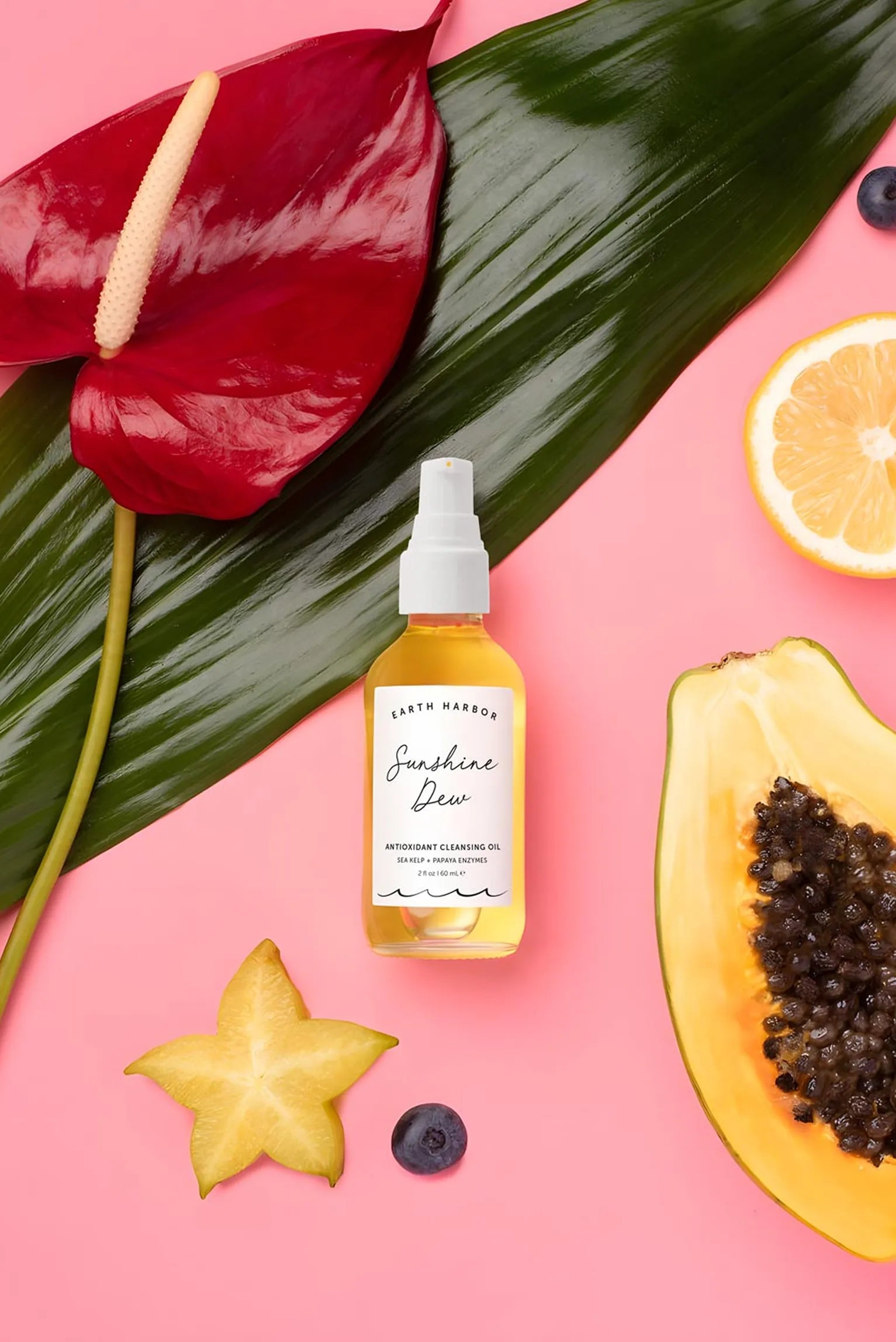Earth Harbor Sunshine Dew bottle on pink
background with palm leaves, papaya,
starfruit, and orange.​