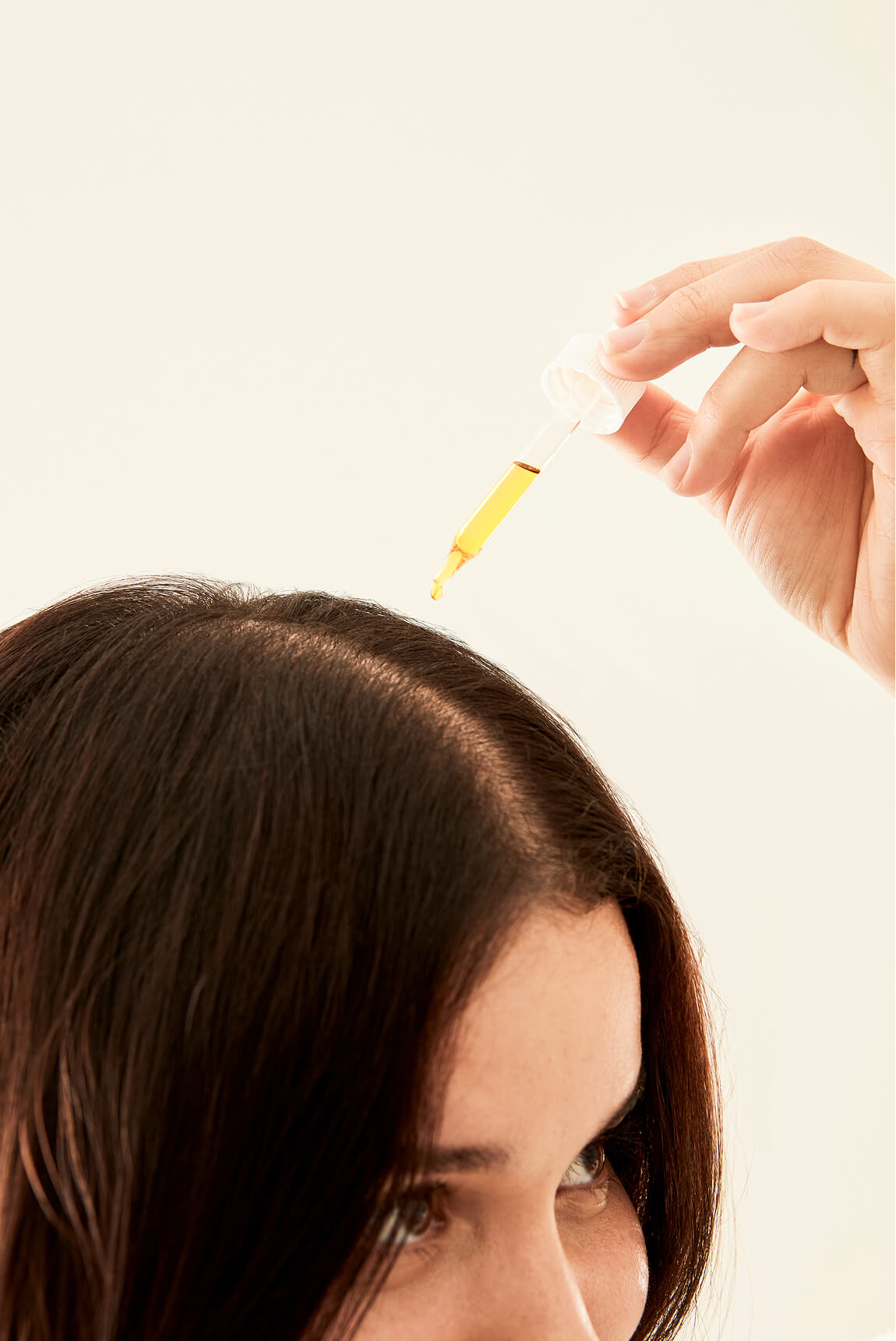 Hand using a dropper to apply golden hair
oil directly onto a woman's scalp parting.​