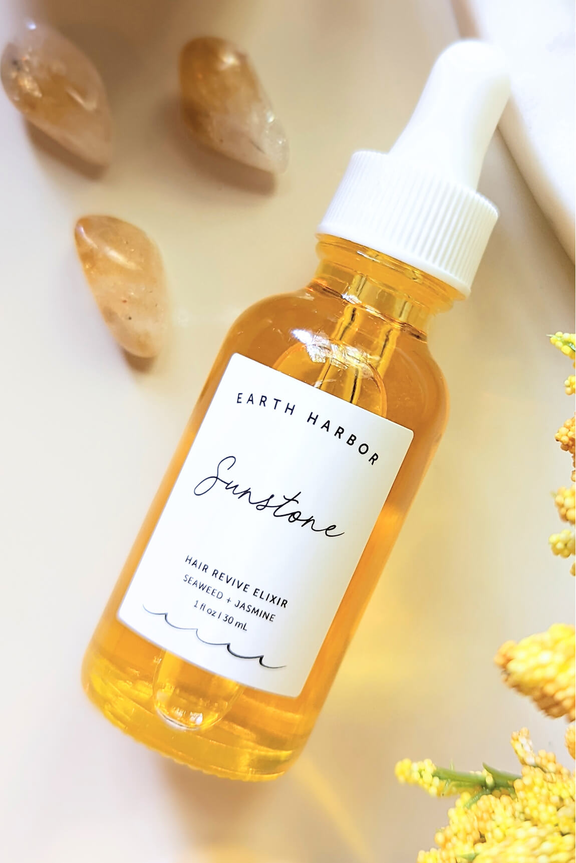 Dropper bottle of Earth Harbor Sunstone
hair elixir next to polished crystals and
yellow flowers.​