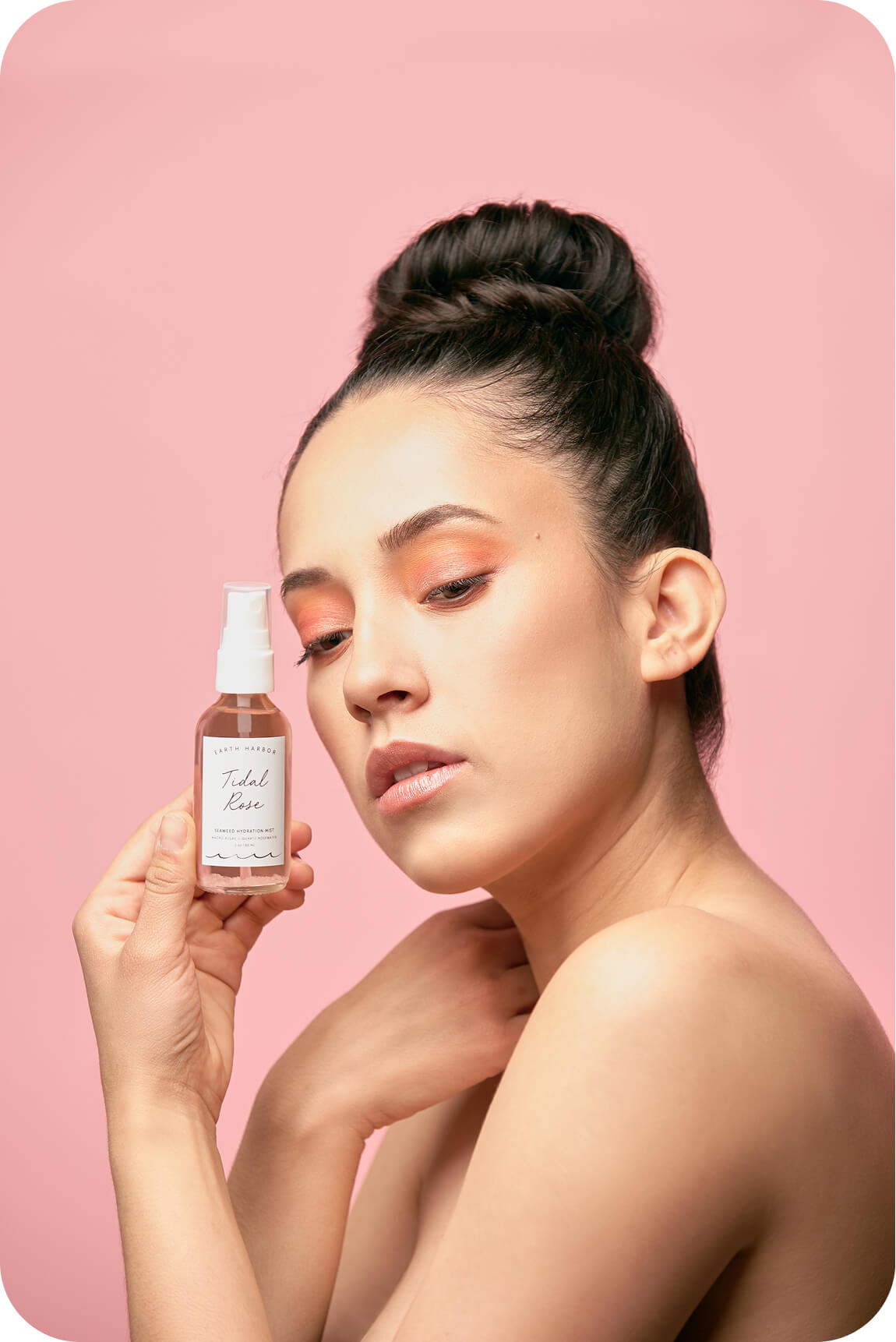 Woman with her hair in a high bun holding
up a small bottle of Earth Harbor Tidal
Rose mist against a pink backdrop.​