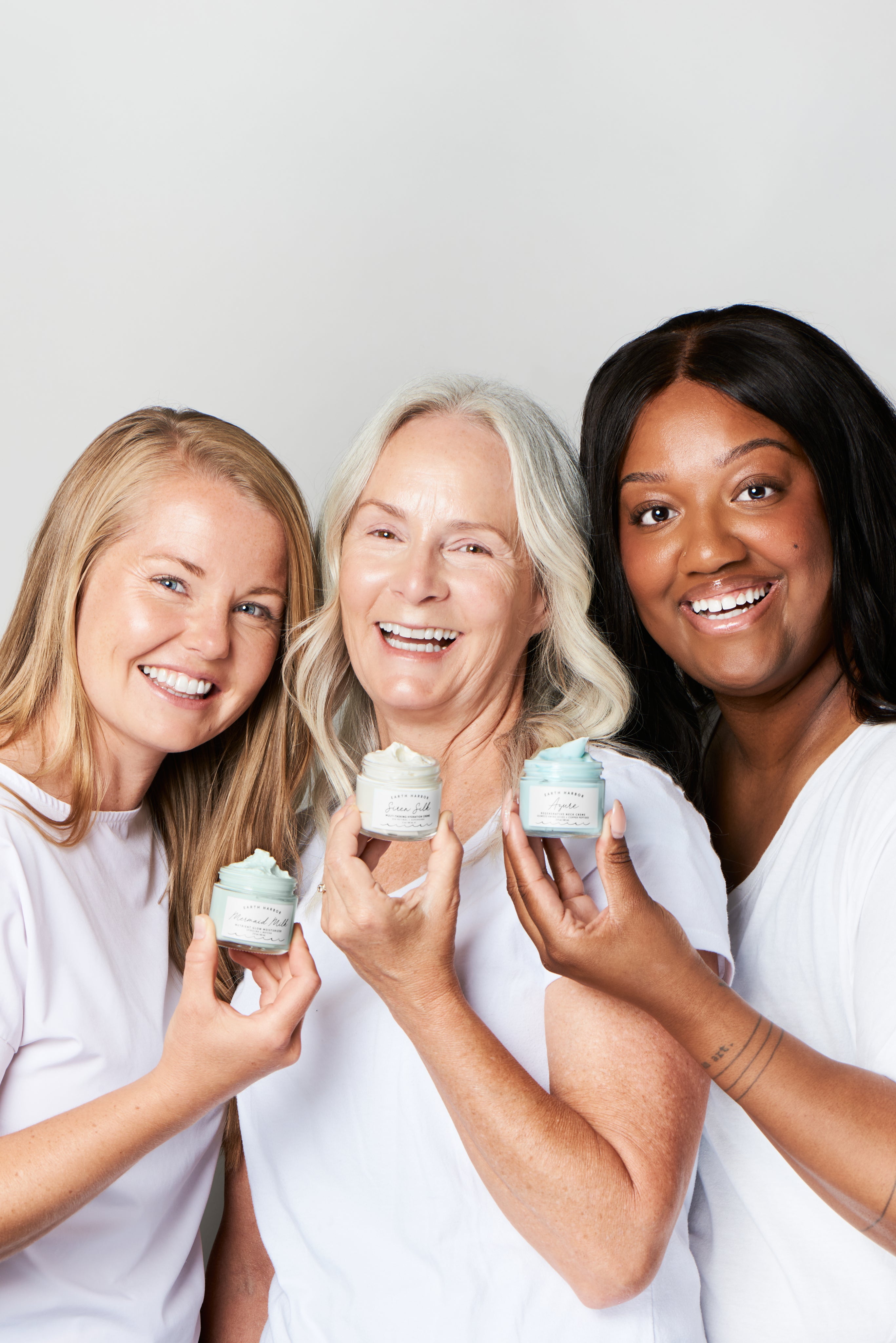 Three women of different ages smiling
and holding skincare jars, showcasing products
together.​